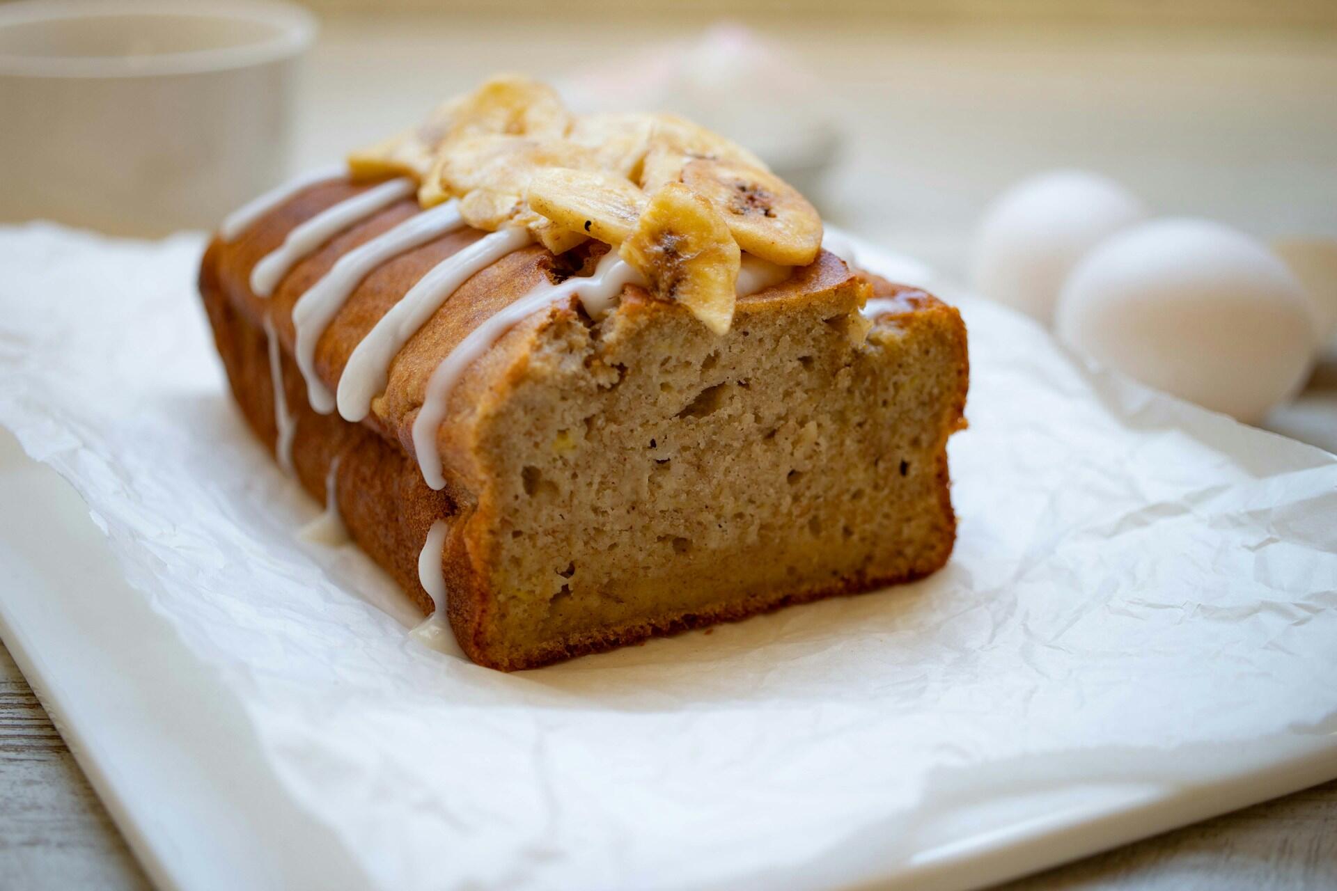 A sliced loaf of banana bread topped with banana slices and drizzled with white icing rests on parchment paper on a plate. 