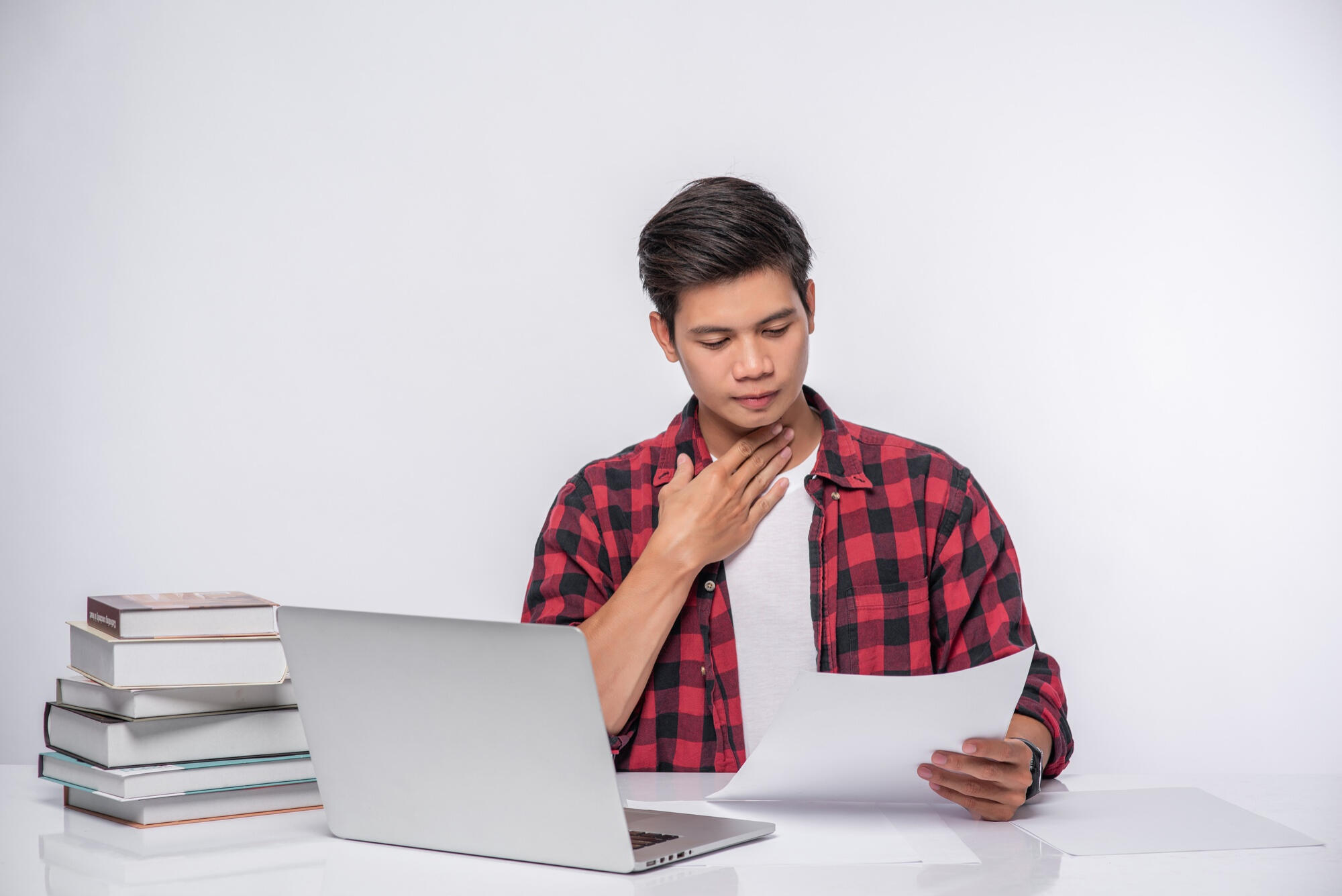 A man in a red plaid shirt touches his throat while reading papers beside a laptop and stack of books.