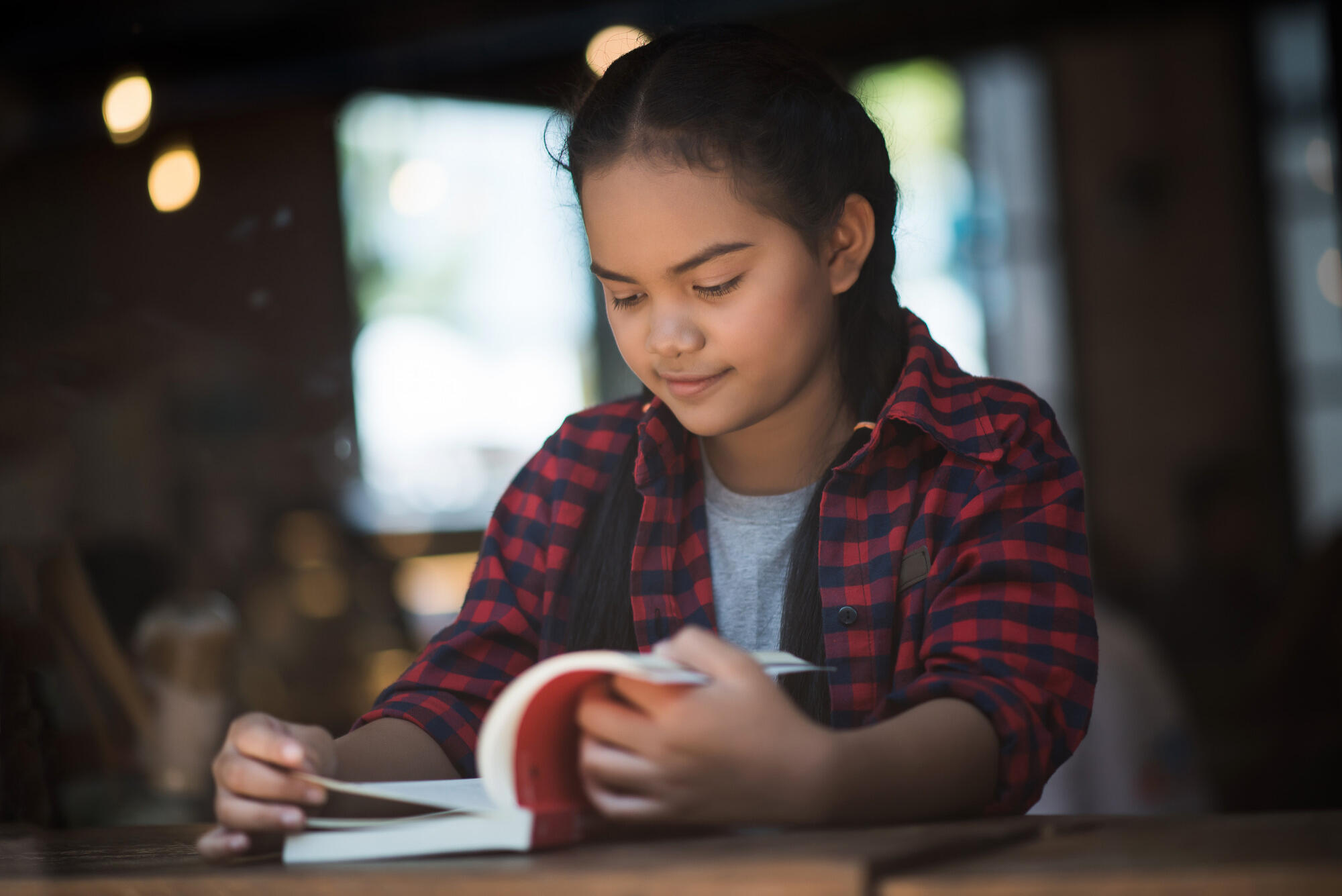 A girl sits at a table, engrossed in reading a book, with a focused expression on her face.