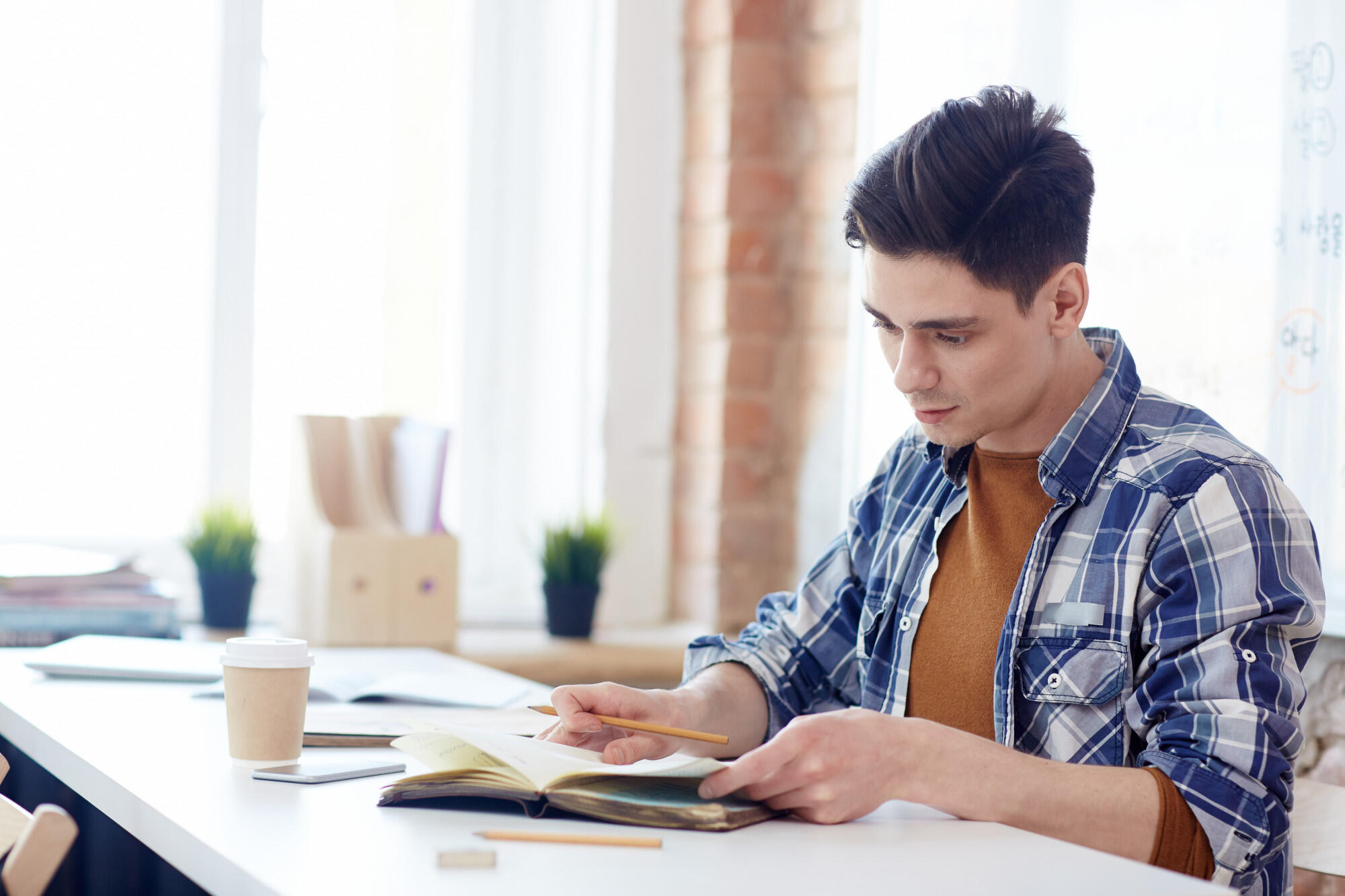 Young man studying an open book and writing with a pencil at a bright desk.