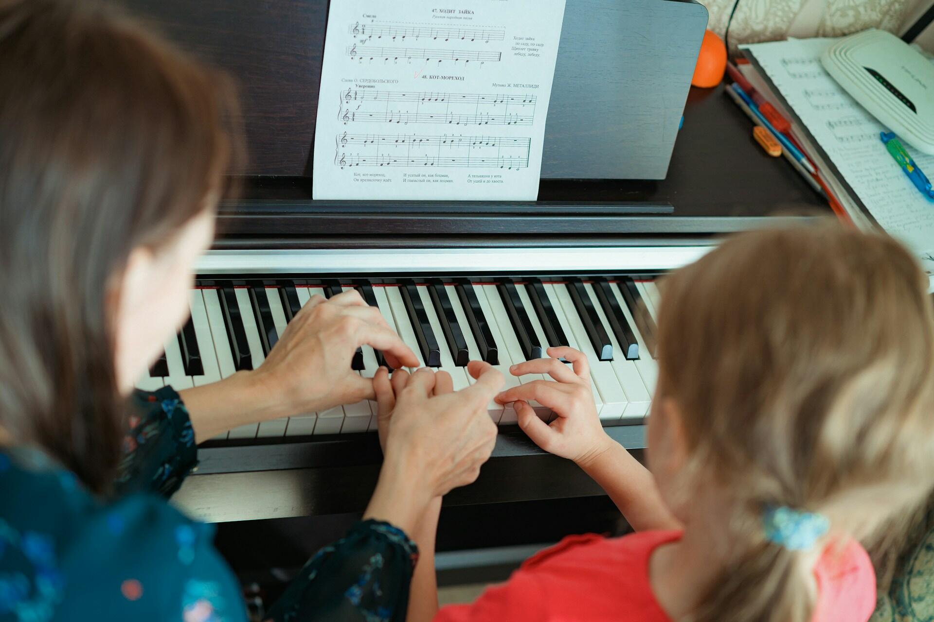 A woman guides a child at a piano, hands together on the keys. Sheet music is on the stand, suggesting a piano lesson. The setting is warm and educational.