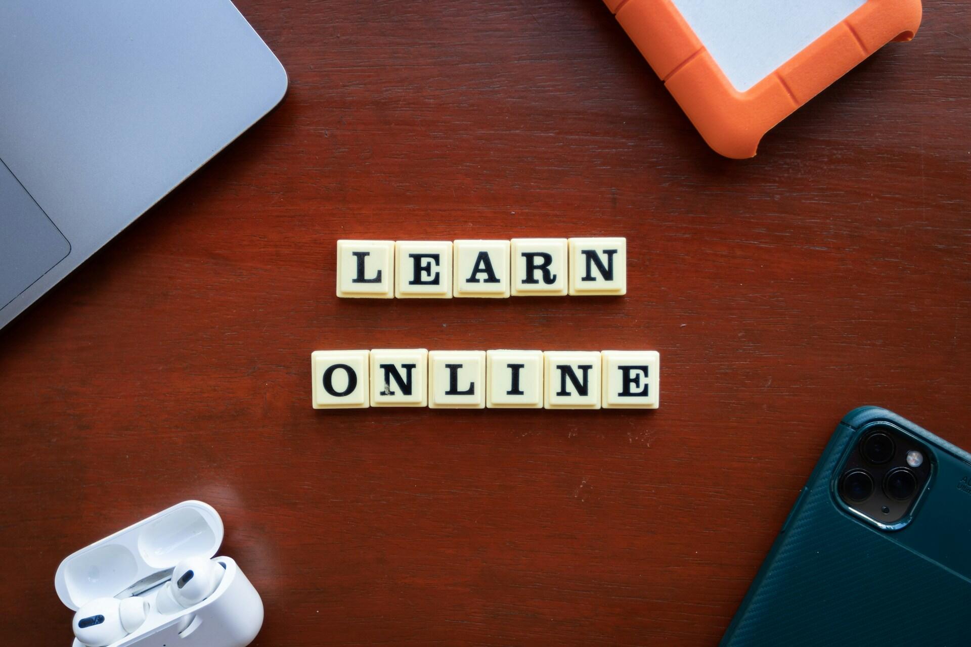 Tiles spell "Learn Online" on a wooden table surrounded by a laptop, tablet, smartphone, and AirPods, conveying a modern, digital learning environment.