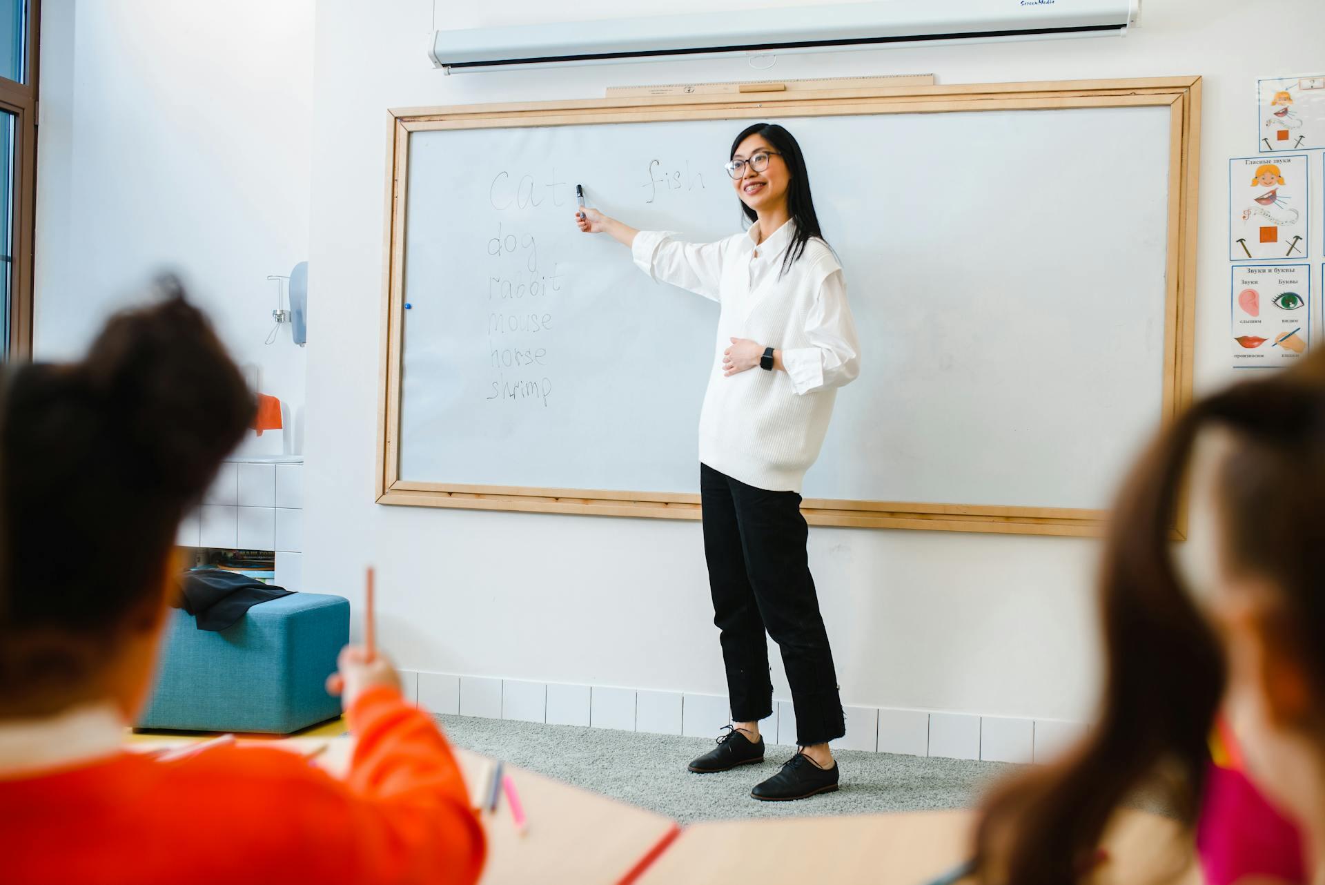 A teacher in a classroom points to a whiteboard with English words written on it. Smiling students in the foreground focus on the lesson.