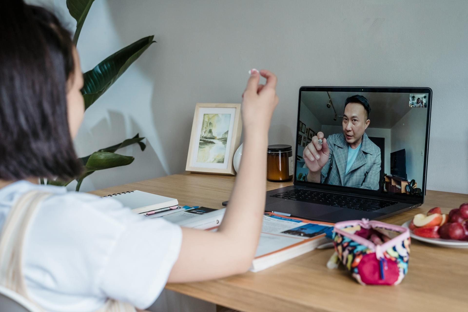 A woman is sitting at a desk, attentively listening to a man on a video call on her laptop. The desk has notebooks, a colorful pouch, and fruit. The mood is focused and educational.