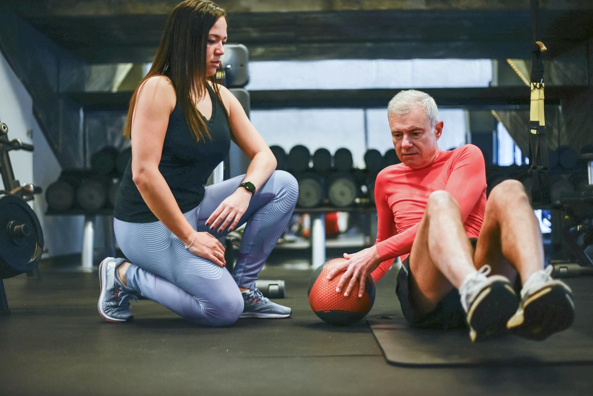 A woman kneels next to an older man in a gym. He is performing a Russian twist with an orange medicine ball. The setting is focused and supportive.