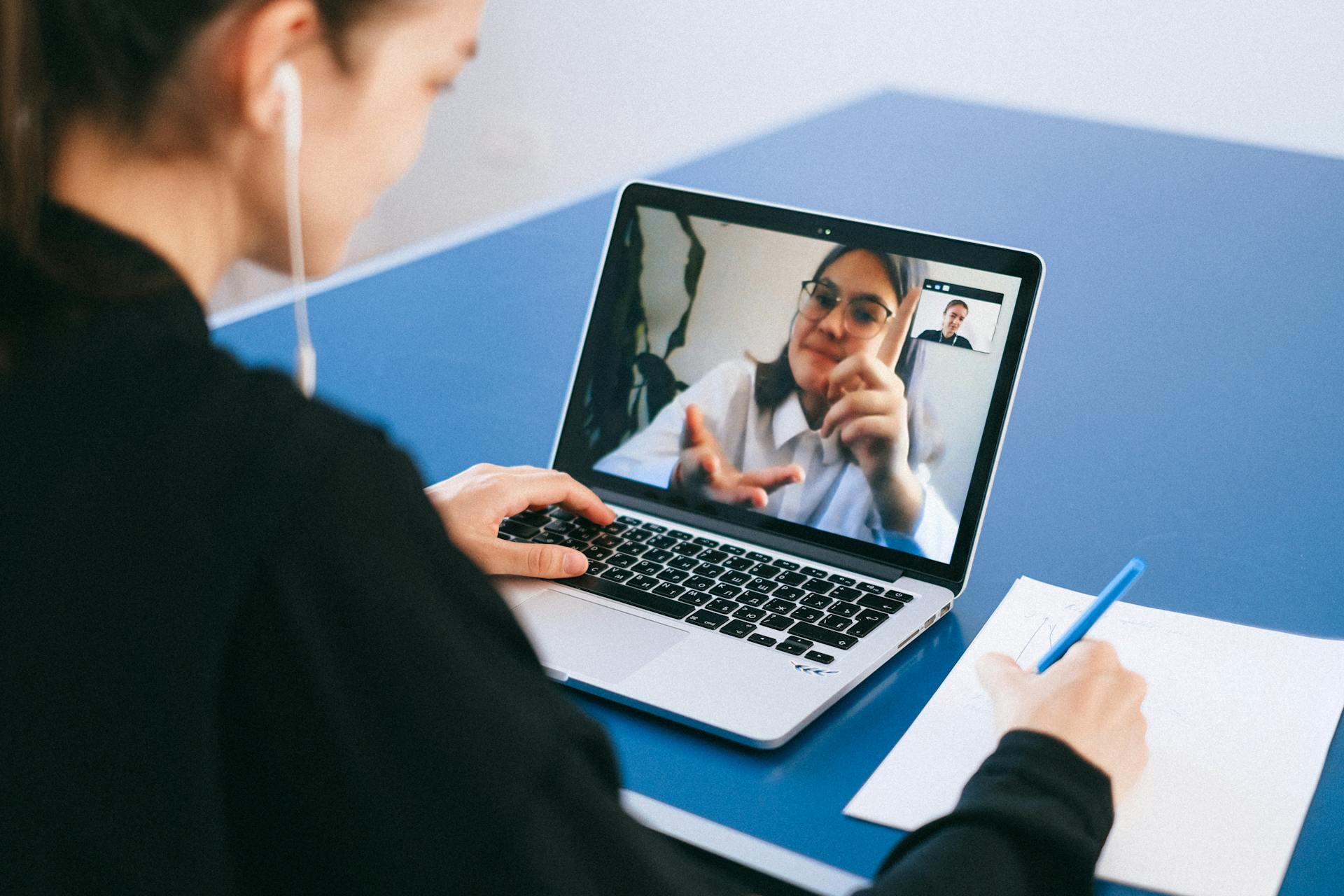 A woman with earbuds participates in a video call on a laptop, engaging attentively with another woman on the screen. She takes notes with a pen.