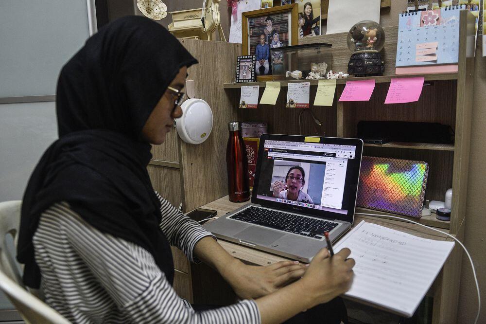 Student in hijab attending online class via laptop while taking handwritten notes at home study desk