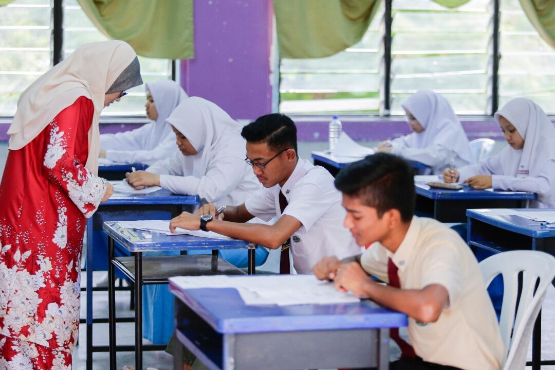 Students taking an exam at desks in a classroom with a teacher in presence