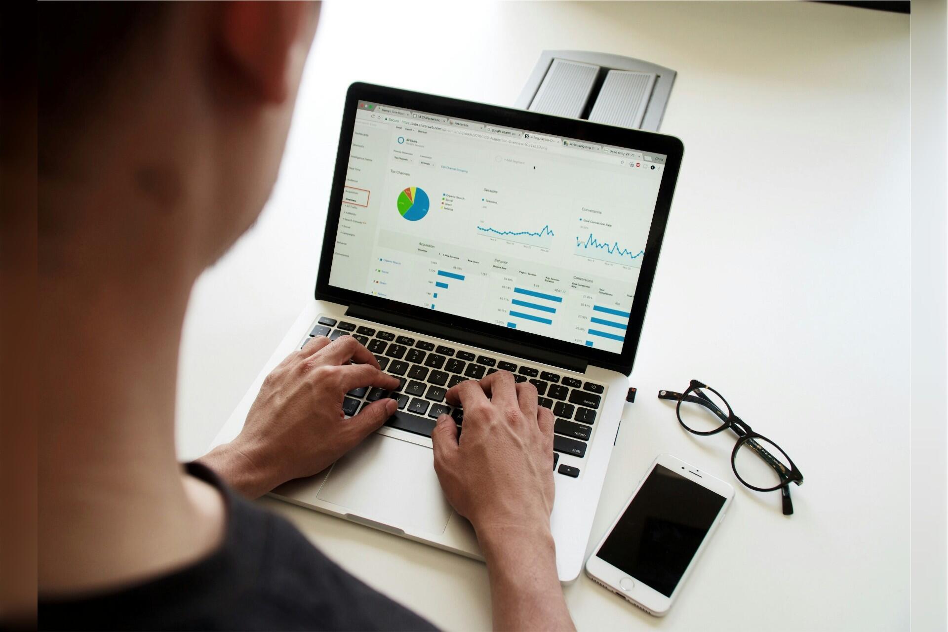 Person analyzing website analytics dashboard with charts and graphs on a laptop, with a smartphone and glasses on the desk.