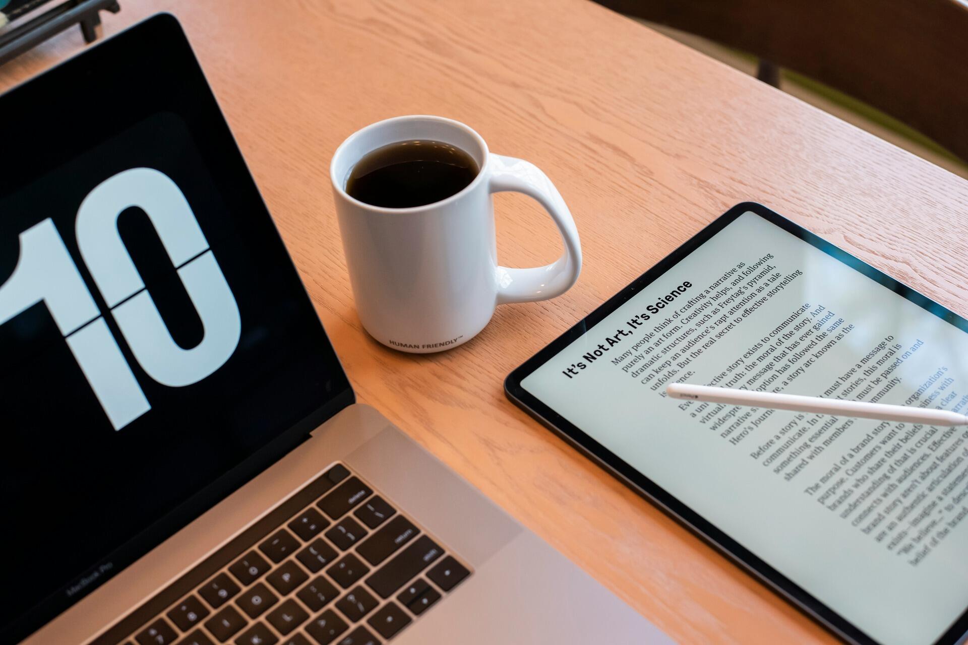 A laptop, a cup of coffee, and a tablet with text displayed on a wooden desk, representing remote studying and digital work.