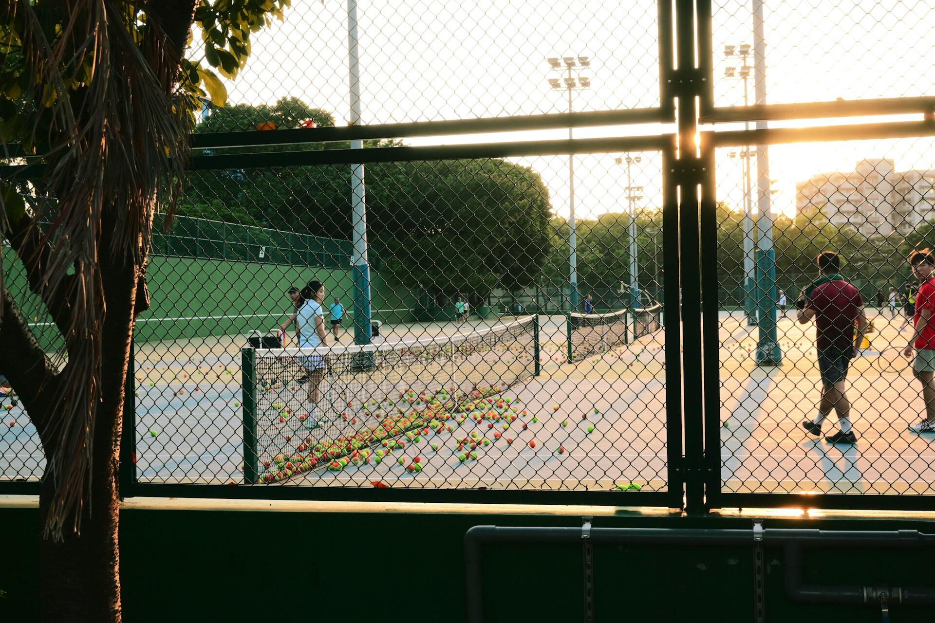 Tennis courts viewed through chain-link fencing with players practicing and colorful training balls scattered on the court during golden hour.