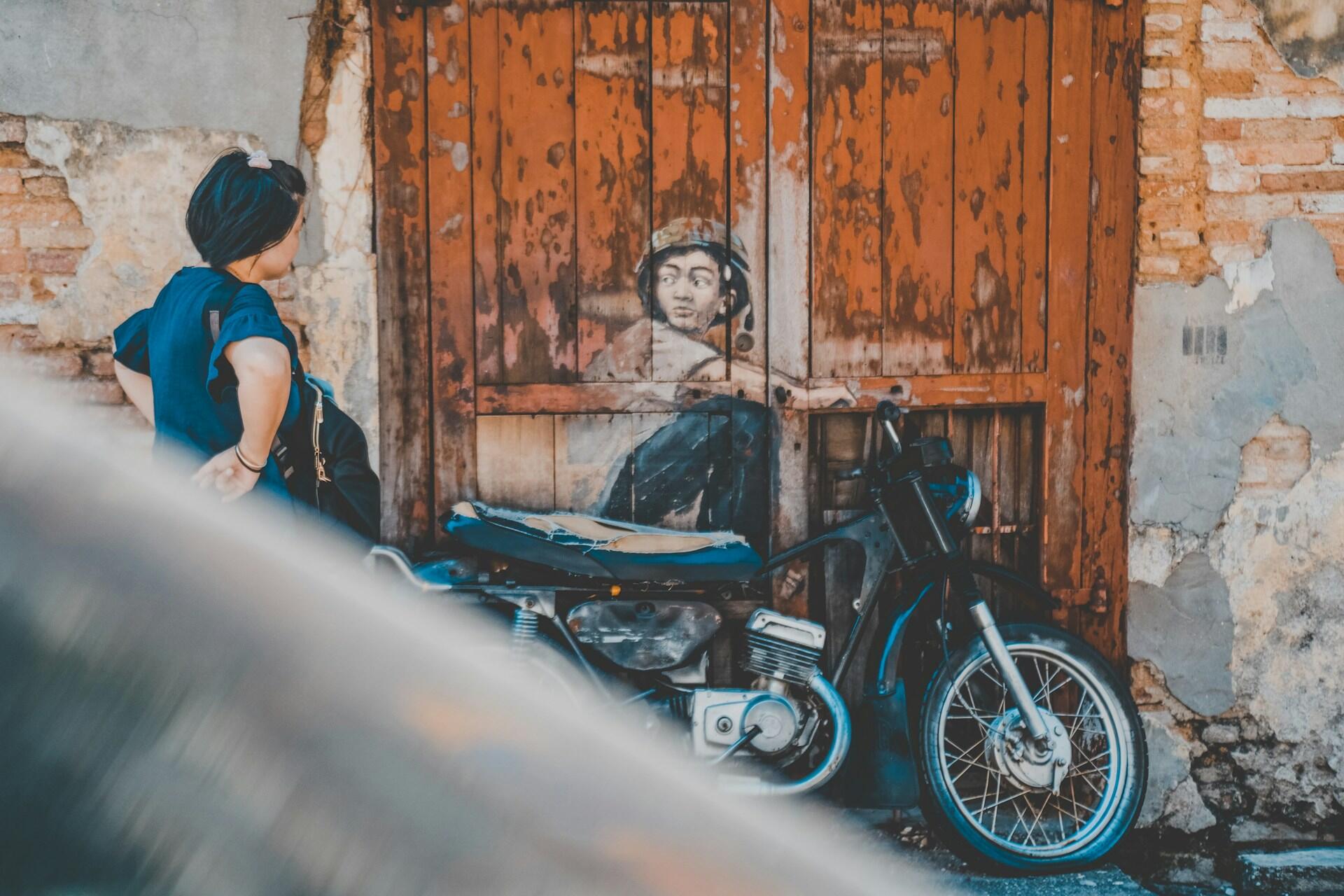 Person sitting on a motorcycle in front of weathered orange wooden doors and a wall with street art.