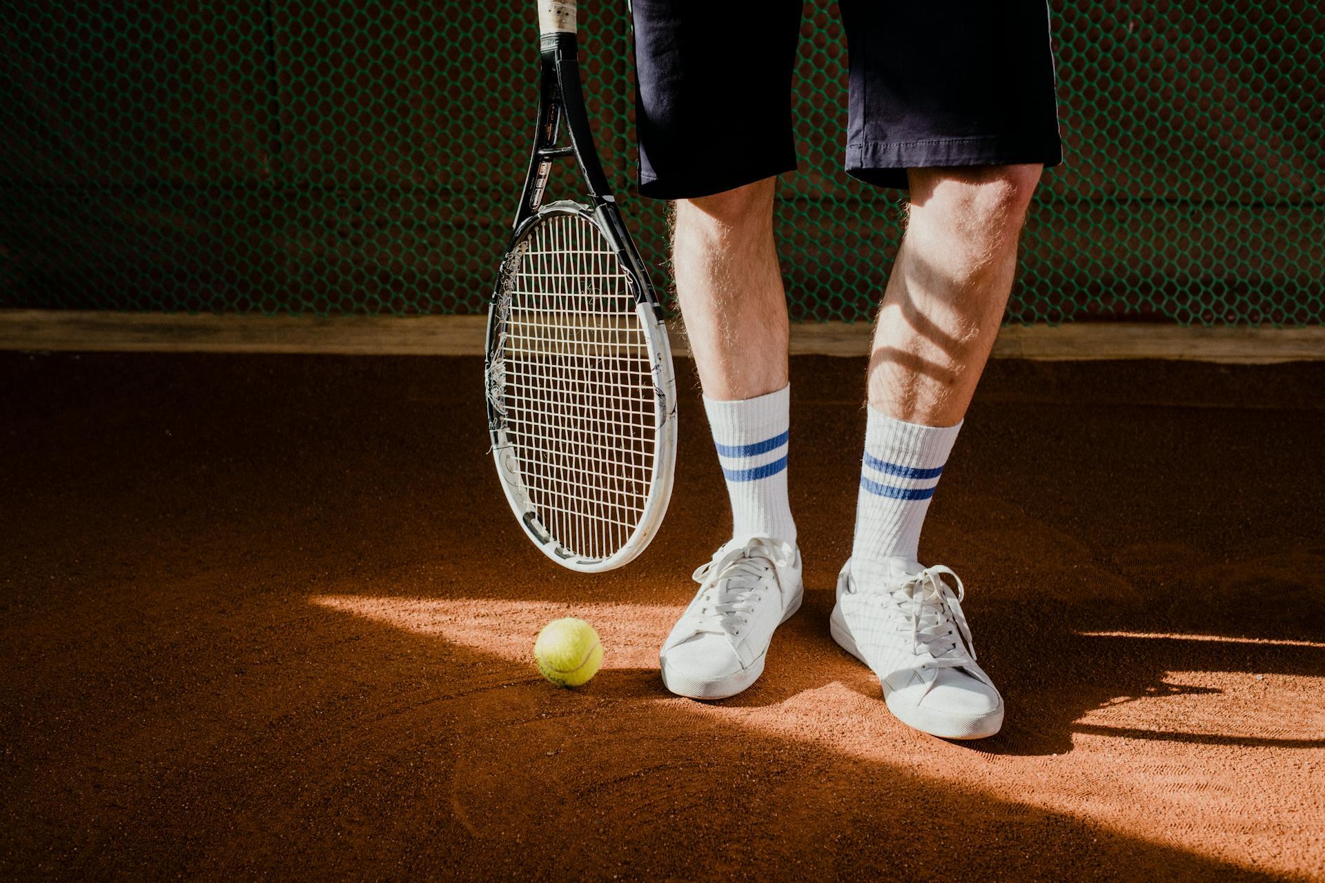A Person Wearing White Sneakers holding a racquet and a yellow ball on the surface of a clay court.