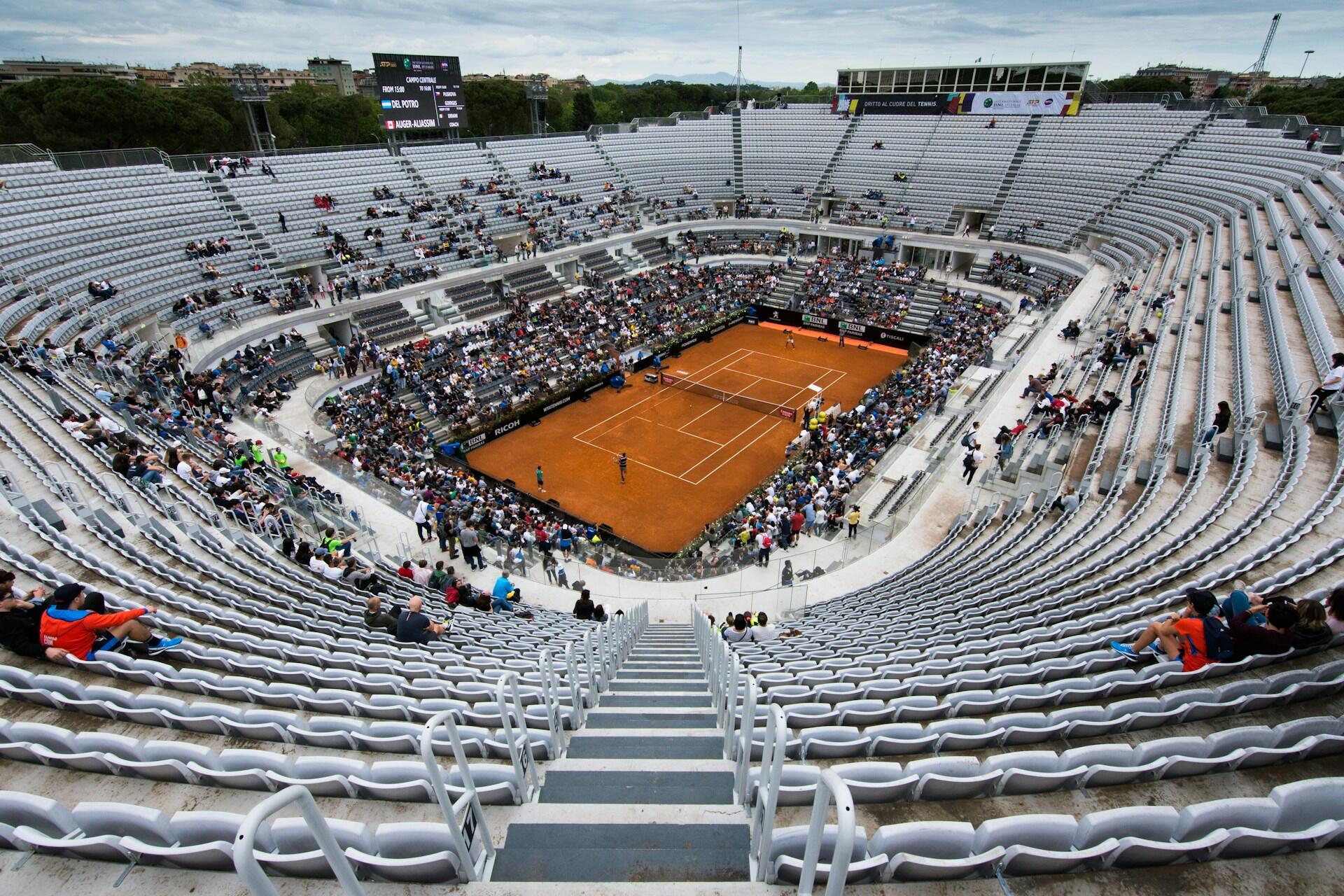 Large tennis stadium with clay court, partially filled stands, and spectators watching a match.