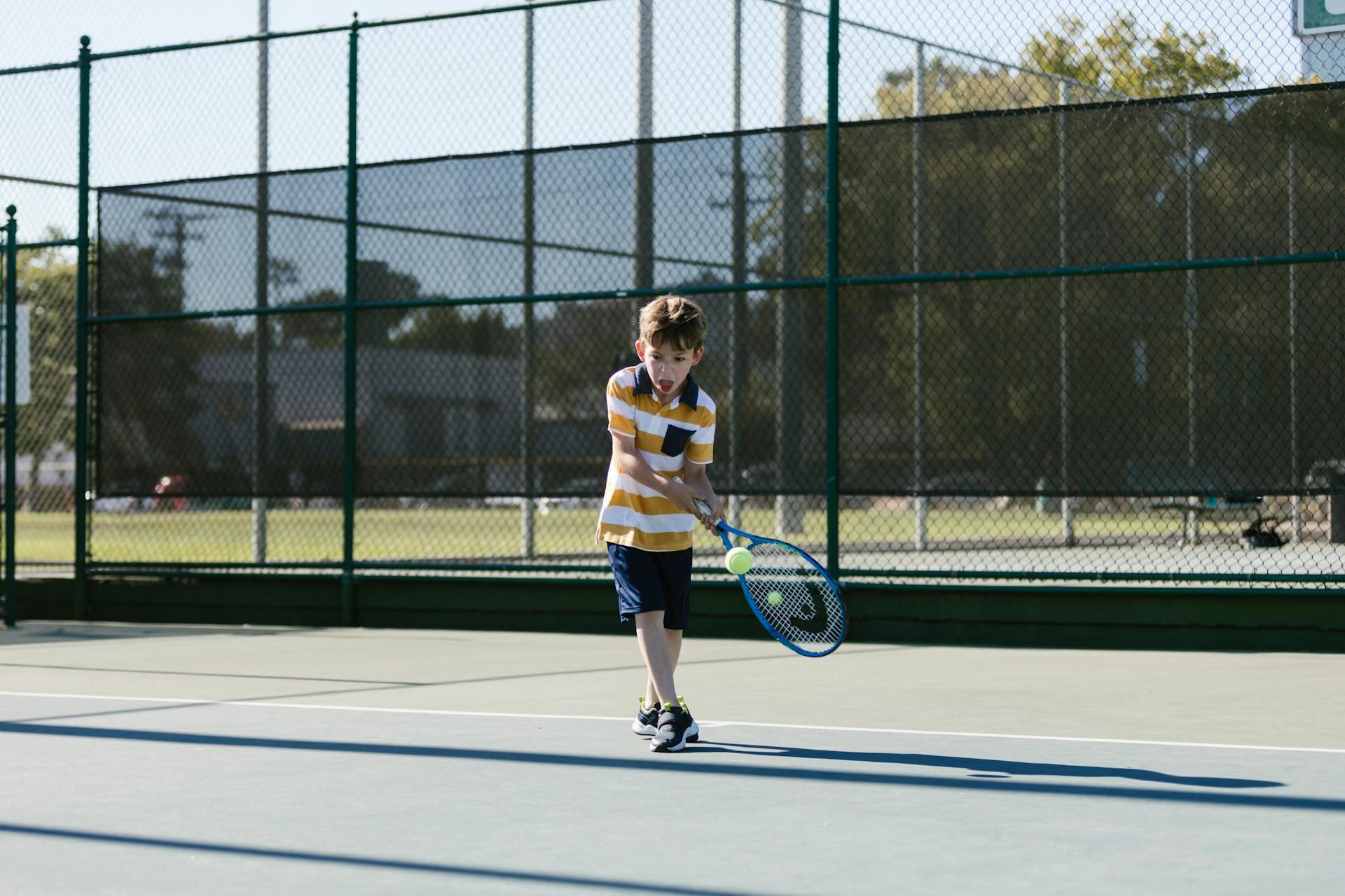 Young boy in a yellow and white striped shirt playing tennis on an outdoor court surrounded by chain-link fencing.