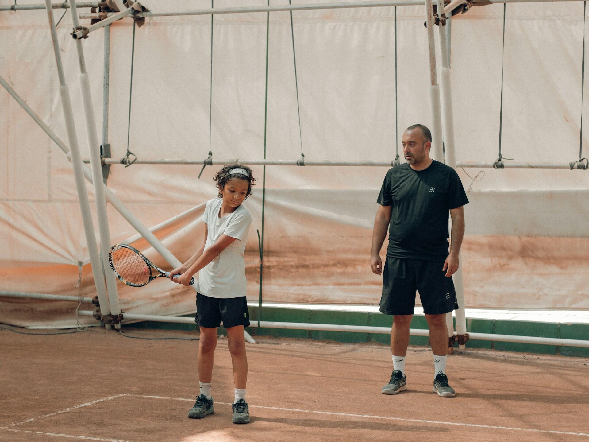 Child learning tennis with adult coach on indoor clay court during a tennis lesson.