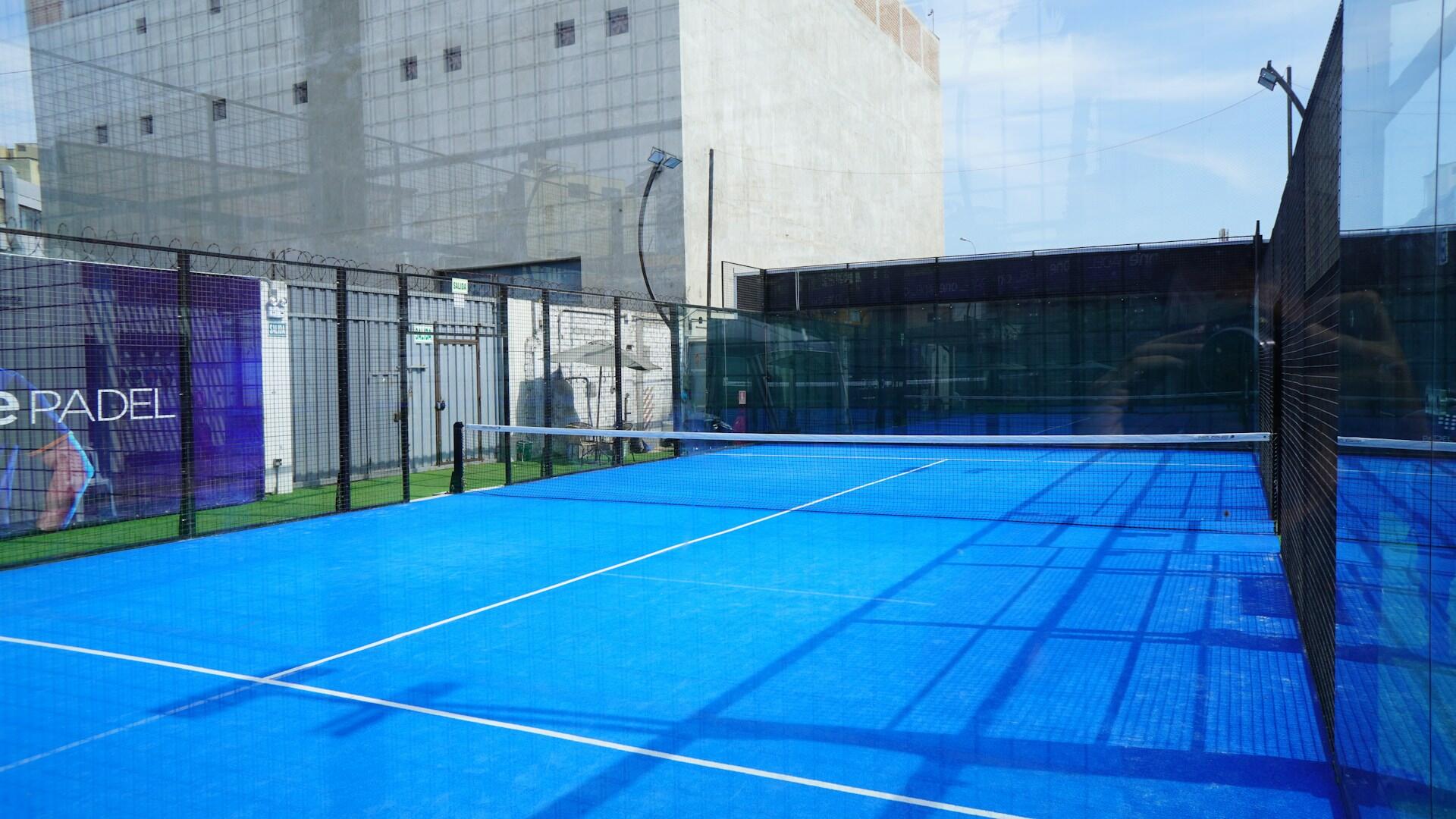 A tennis court with a blue surface surrounded by glass walls and metal mesh fencing with modern buildings in the background.