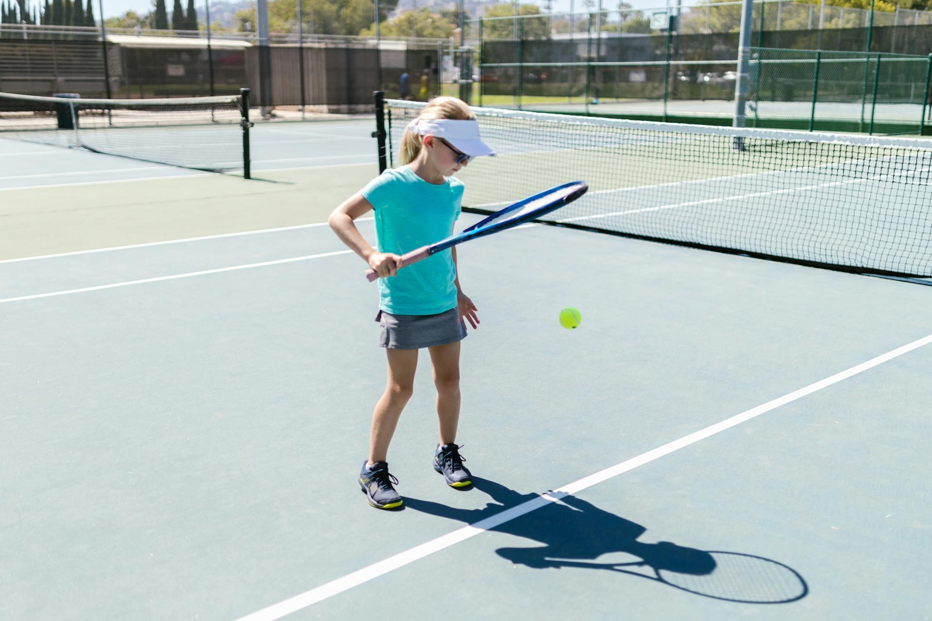 Young girl in a teal shirt and white visor practicing tennis with a racket on an outdoor court.