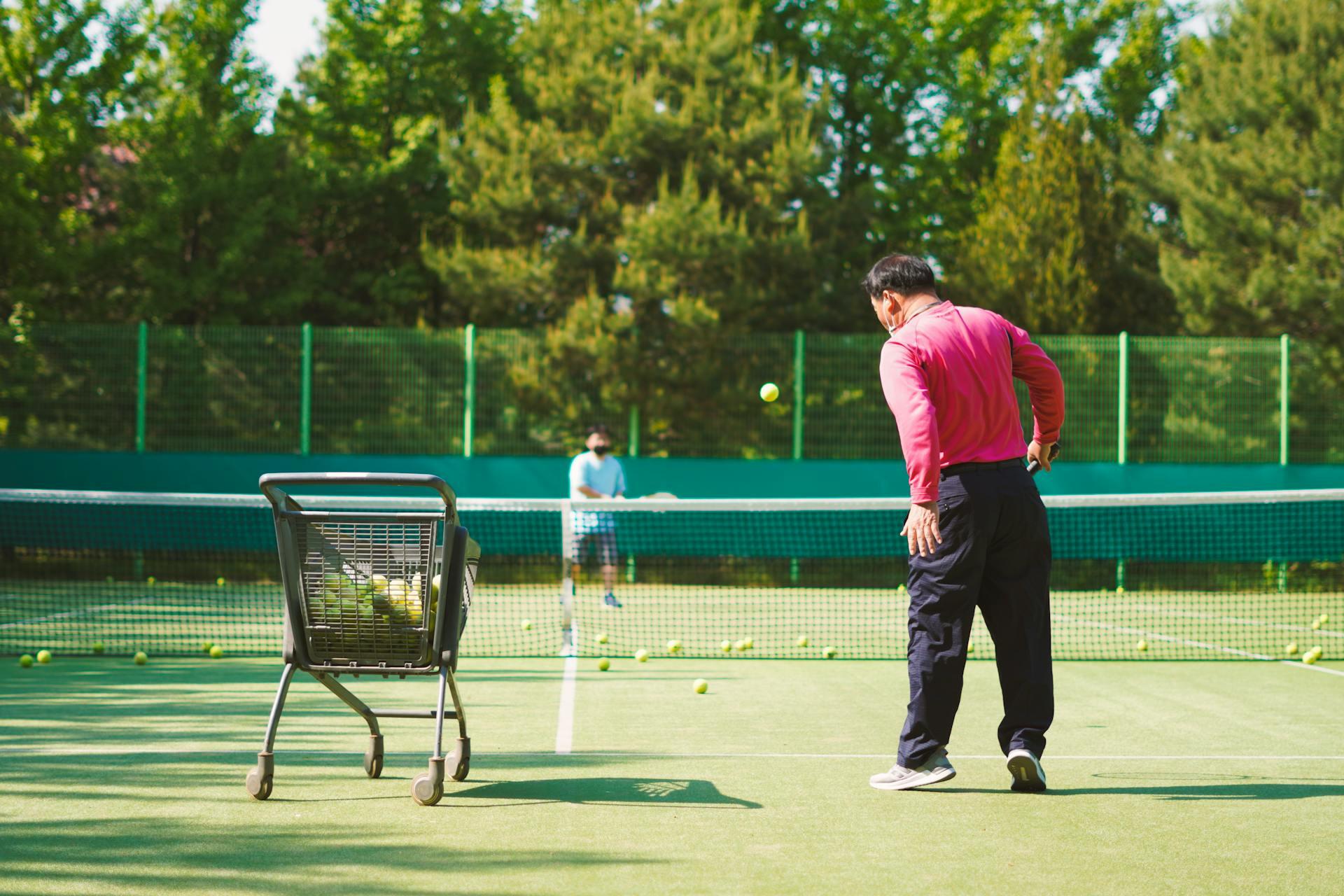 Tennis instructor in a pink shirt standing next to a ball cart on a tennis court with a student in the background.