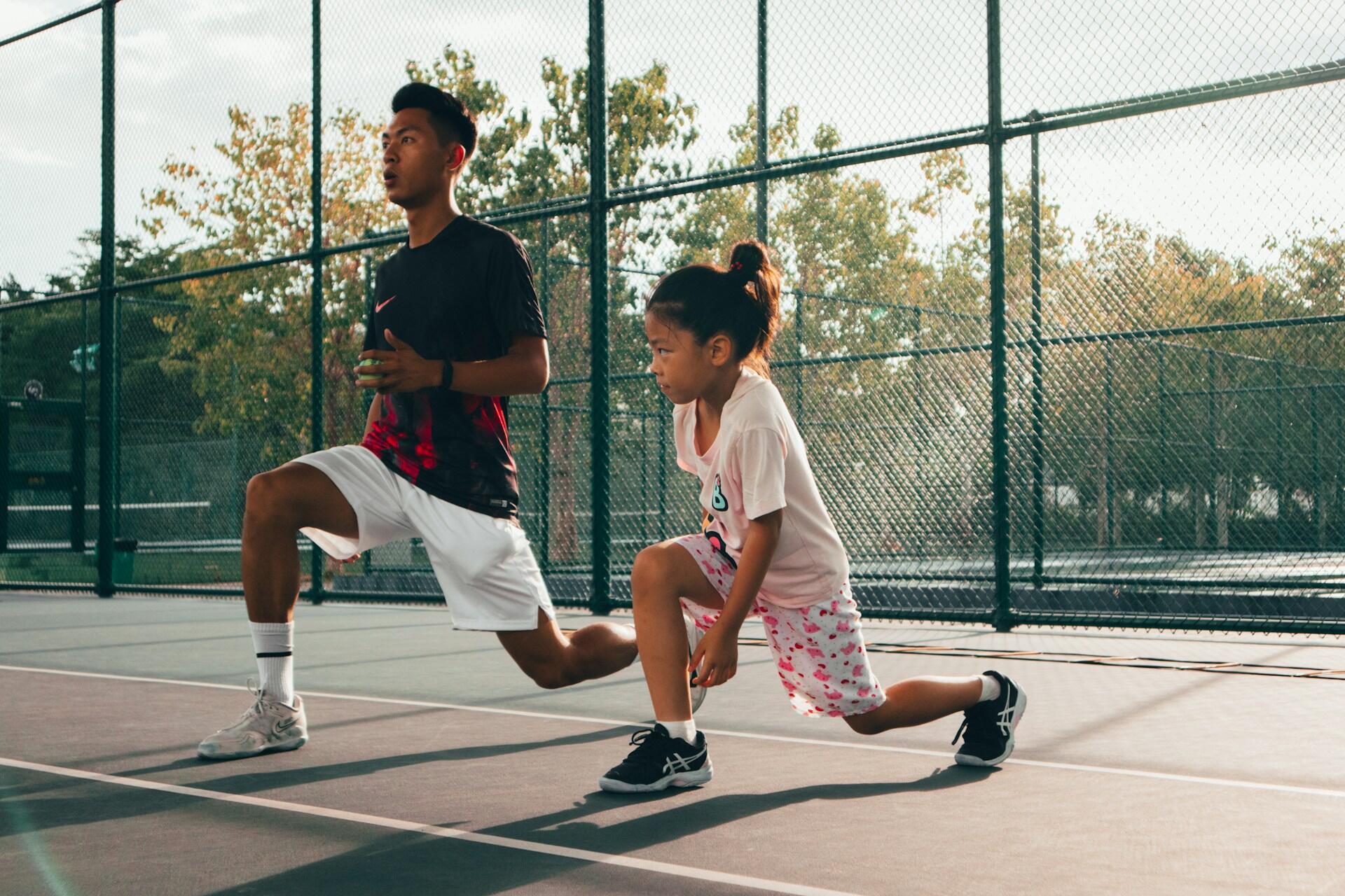 A man and a little girl are seen doing some stretches on the court before playing. 