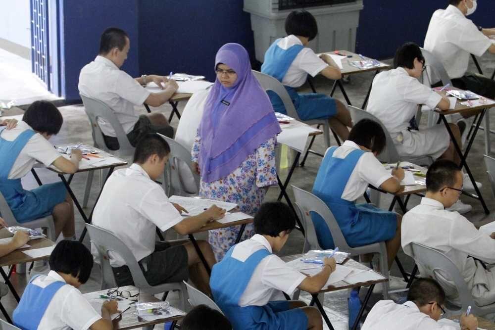 Students in blue uniforms sit at desks in a large exam room, focused on writing. A teacher in a purple headscarf supervises, creating a calm, studious atmosphere.