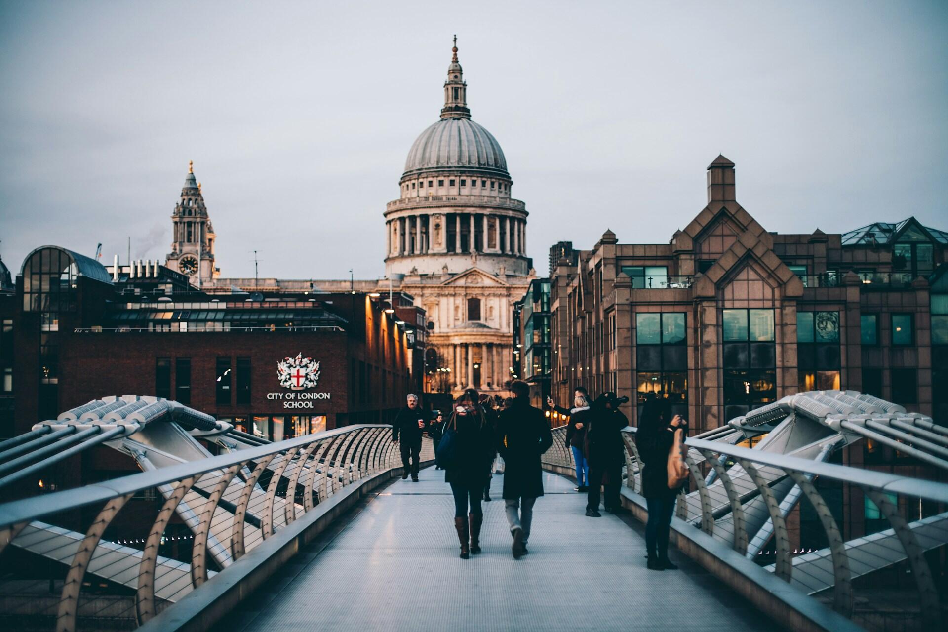 People walk across the Millennium Bridge in London at dusk, with St. Paul's Cathedral in the background, creating a calm urban scene.