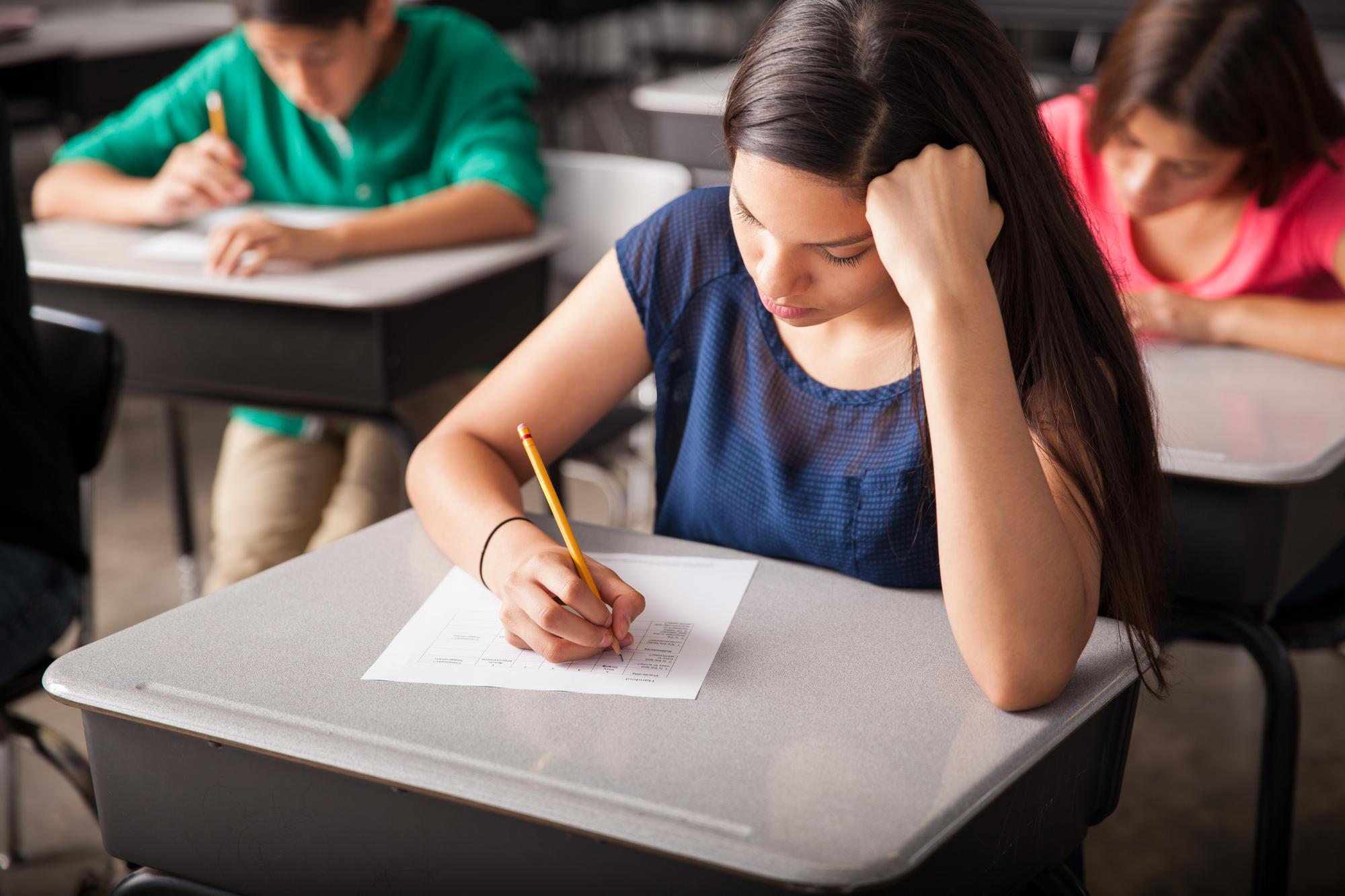 Student concentrating on writing a test or assignment paper in a classroom with peers in the background.