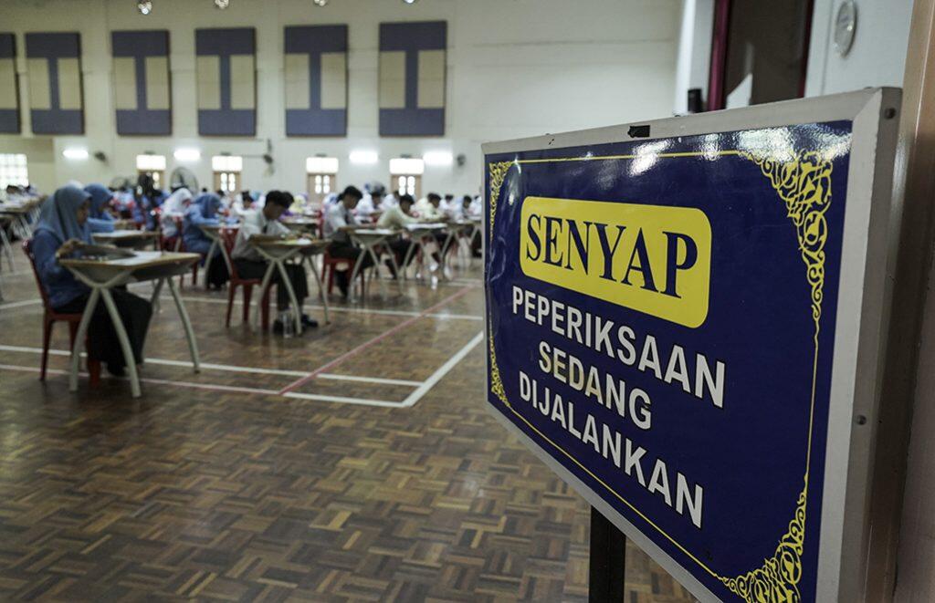 Students sit at desks in a large hall, taking an exam. A sign in the foreground reads "Senyap, Peperiksaan Sedang Dijalankan," urging silence.