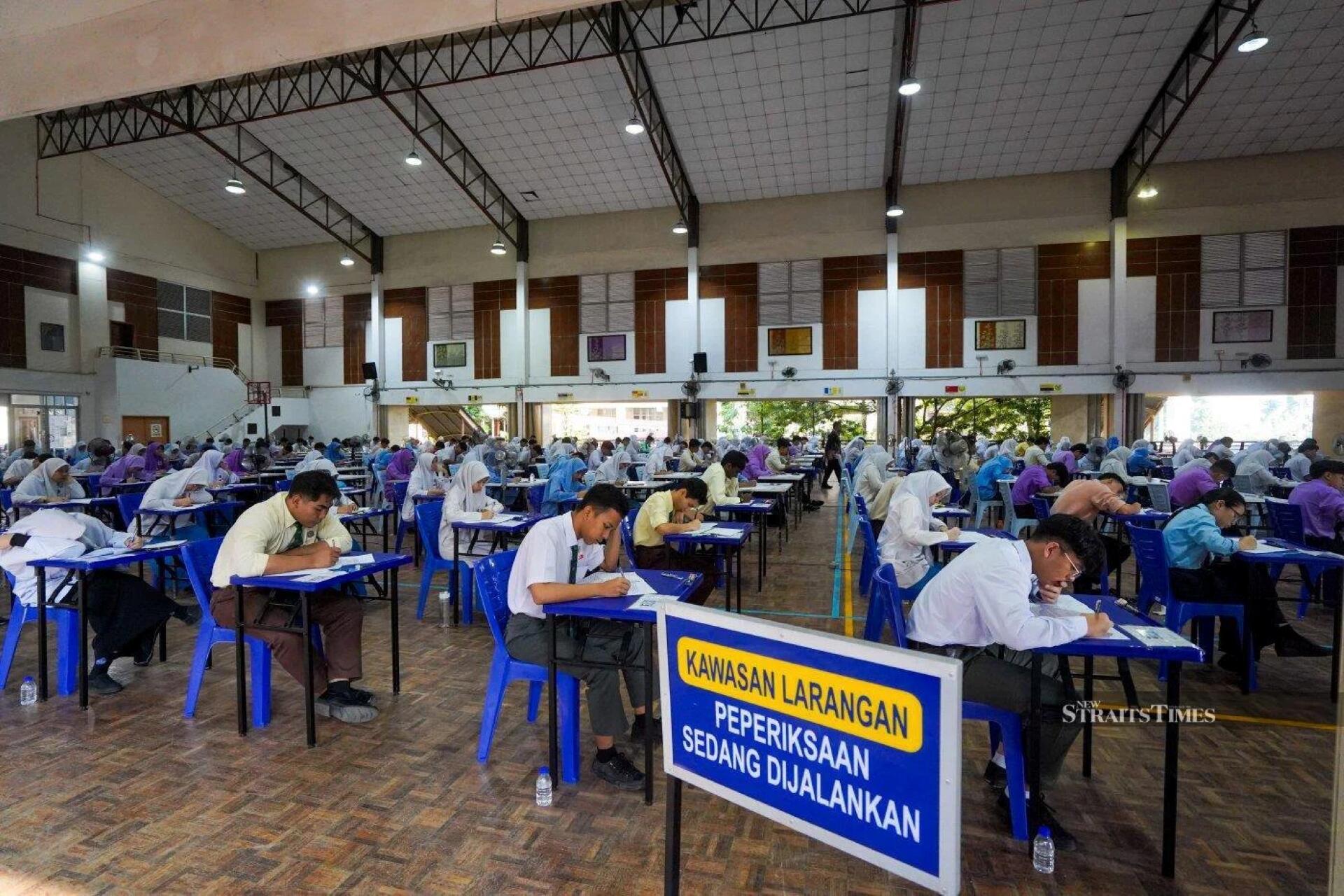 Large hall with students seated separately, focused on writing exams; a sign in Malay indicates exam in progress area.