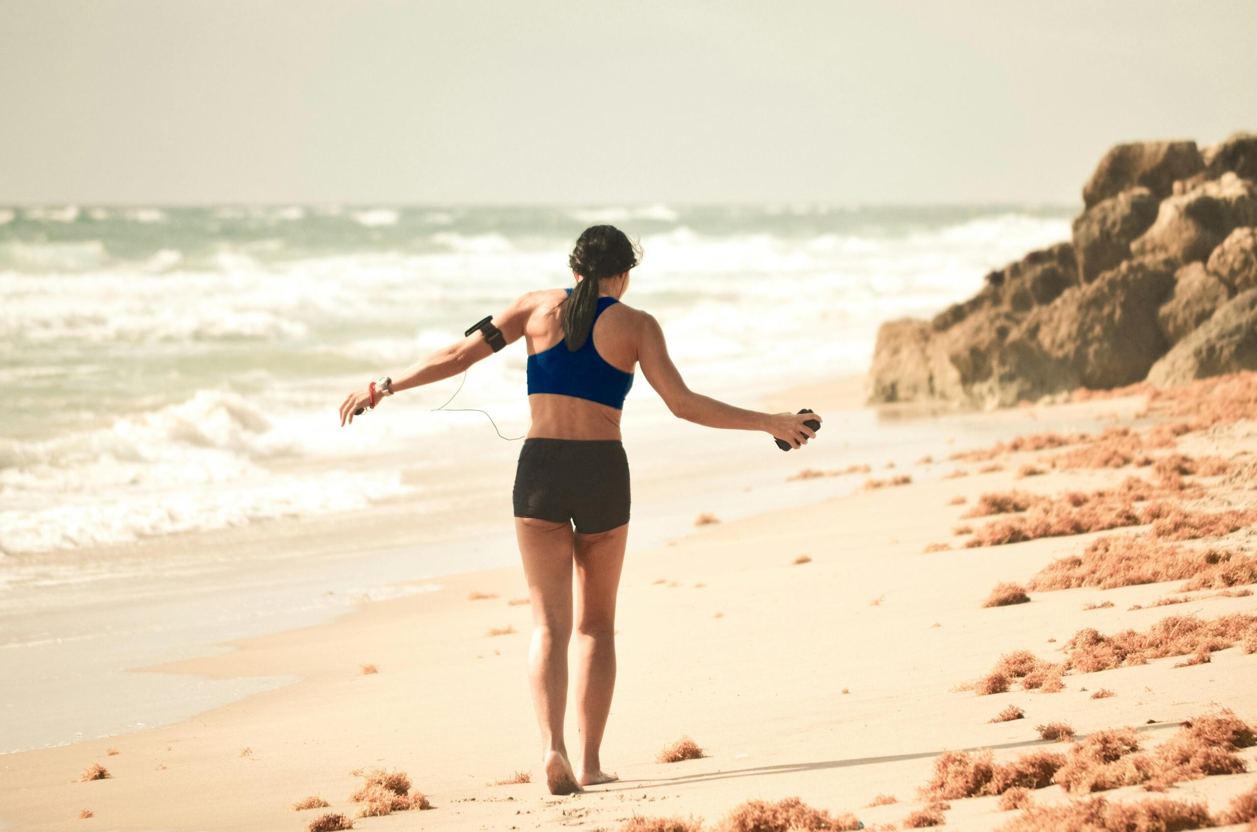Woman jogging barefoot on a beach with waves, rocks, and seaweed in the background.