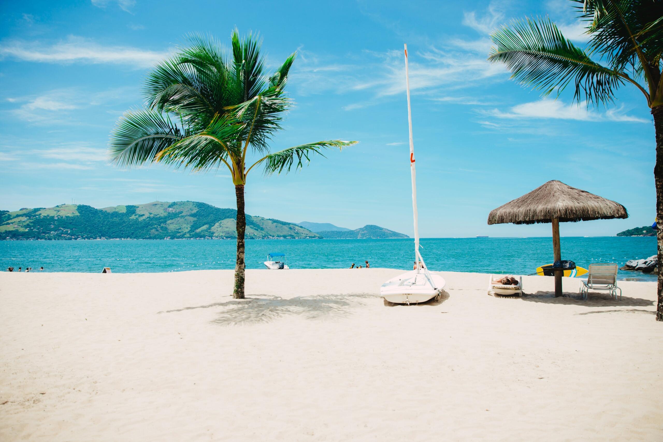 Tropical beach with palm trees, a sailboat on the sand, lounge chairs under a thatched umbrella, and ocean views with distant hills.