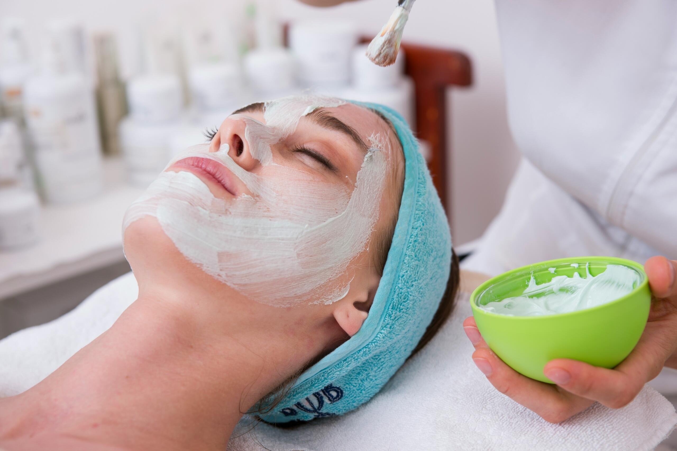 Person receiving a facial mask application at a spa, with a brush applying cream and a headband holding their hair back.
