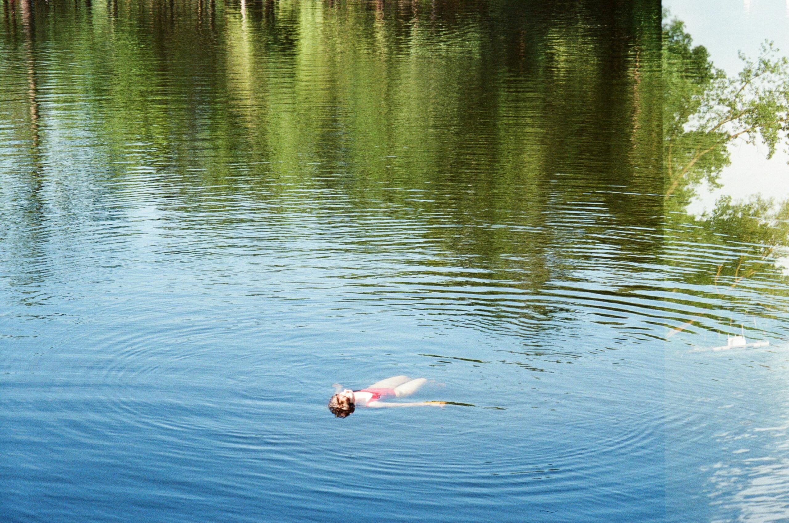 A person floats calmly on their back in a still lake surrounded by reflections of green trees.