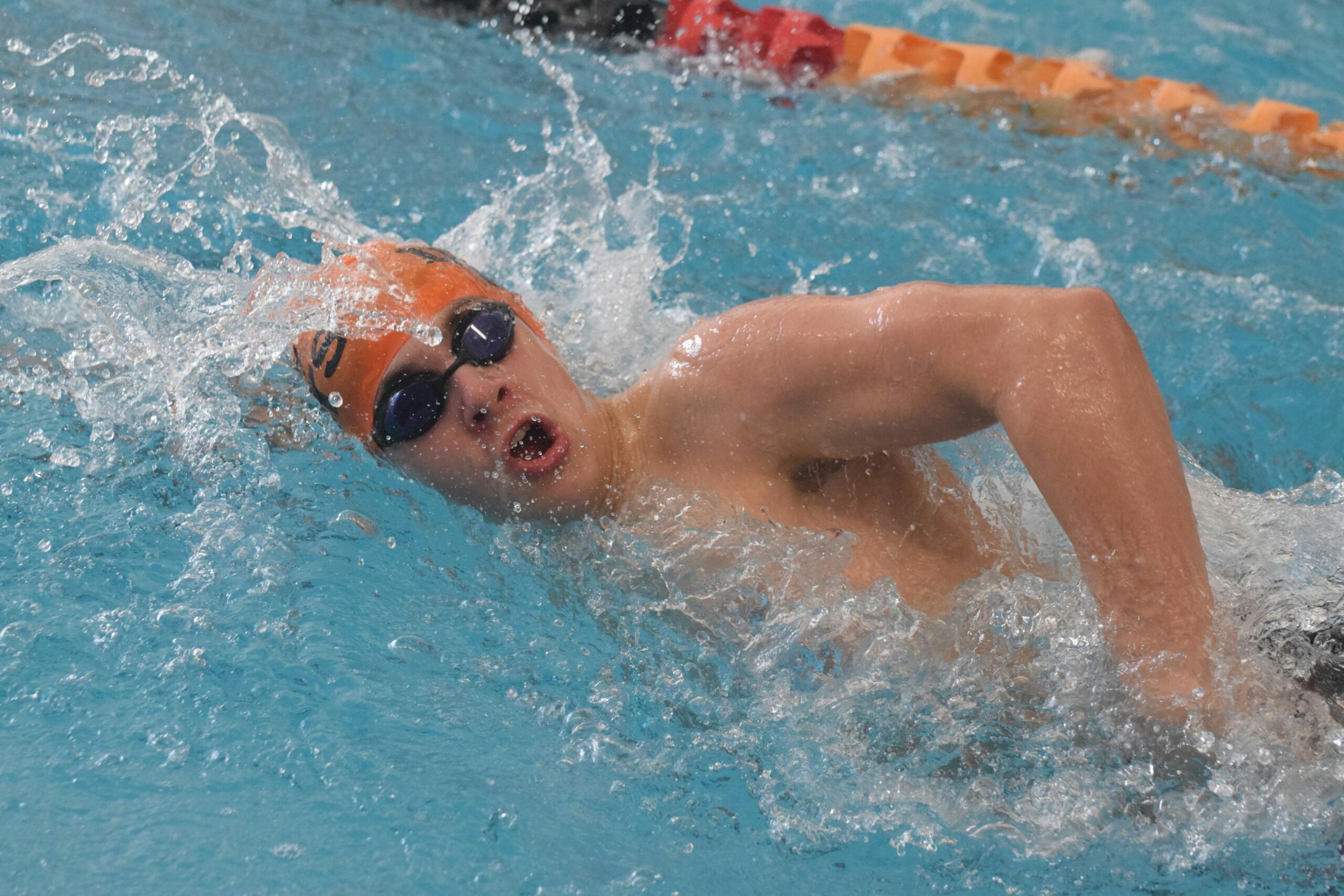 High-energy photo of a male swimmer in an orange cap and goggles taking a breath during a competitive freestyle race, surrounded by water splash.