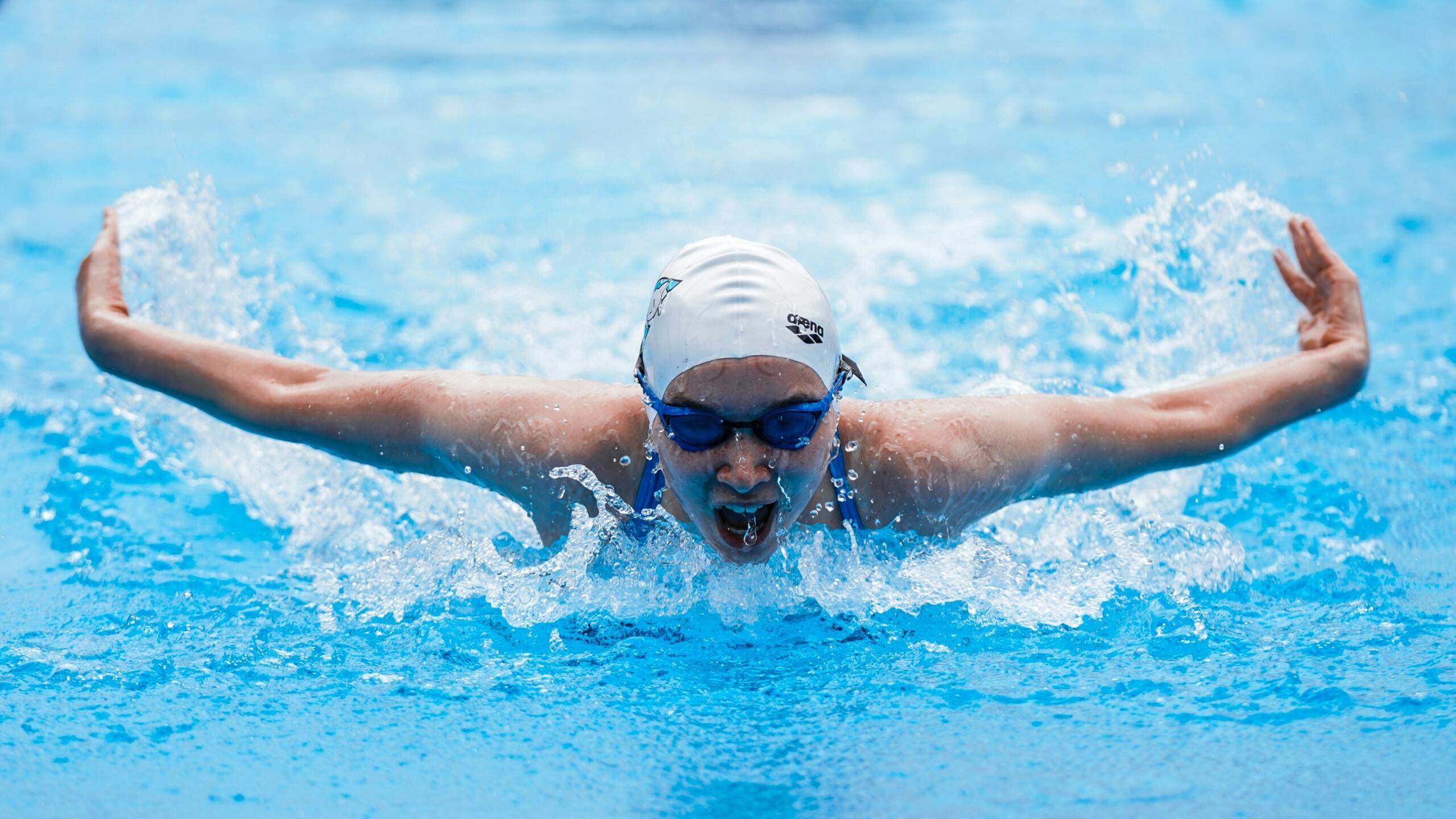 A competitive swimmer performing the butterfly stroke, emerging from the water with a splash, wearing a white cap and blue goggles.