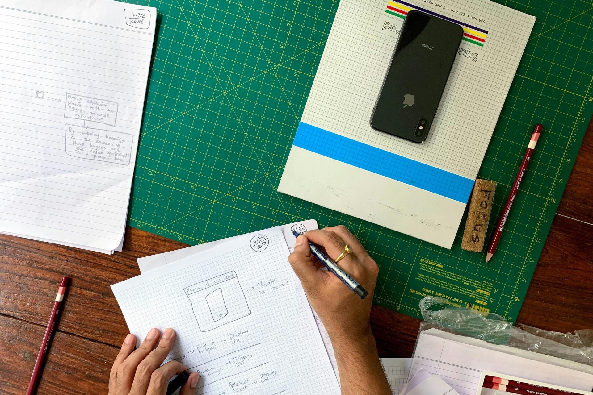 Student writing notes on graph paper at a study desk with stationery, notebooks and a smartphone.