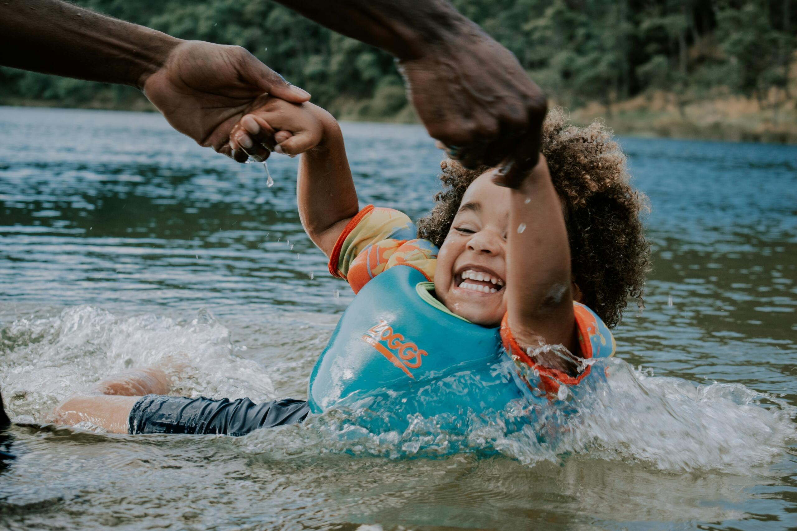 A joyful child wearing a floatation vest splashes in shallow water while an adult holds their hands above the surface.