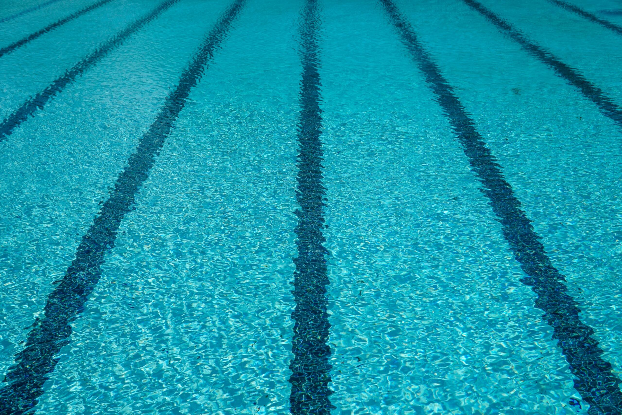 An overhead view of a bright turquoise swimming pool, showing the black lane lines extending diagonally toward the top of the frame.