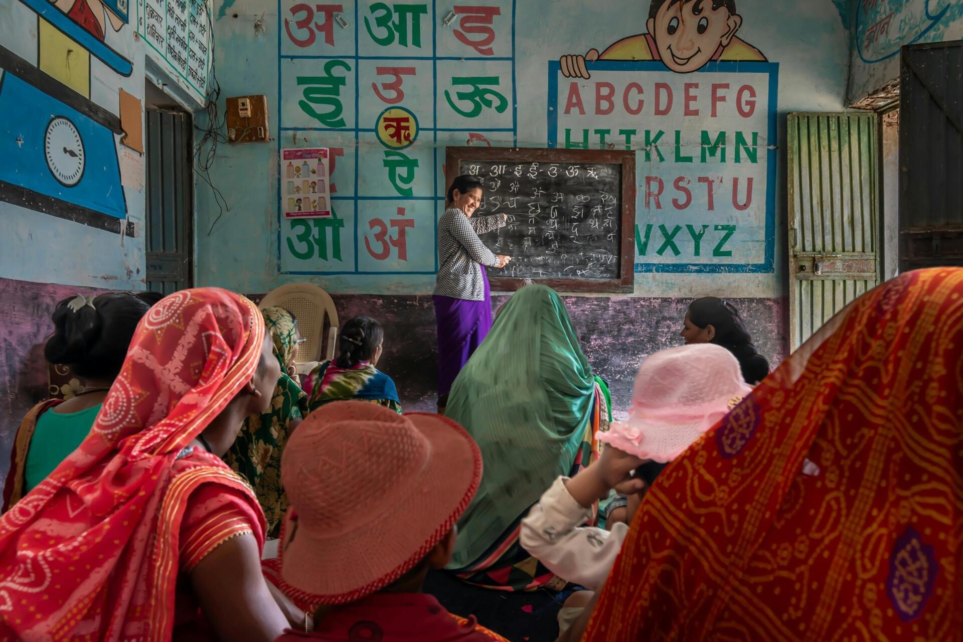eacher conducting a classroom lesson in an educational setting with Hindi and English alphabet charts displayed on the walls as students observe.