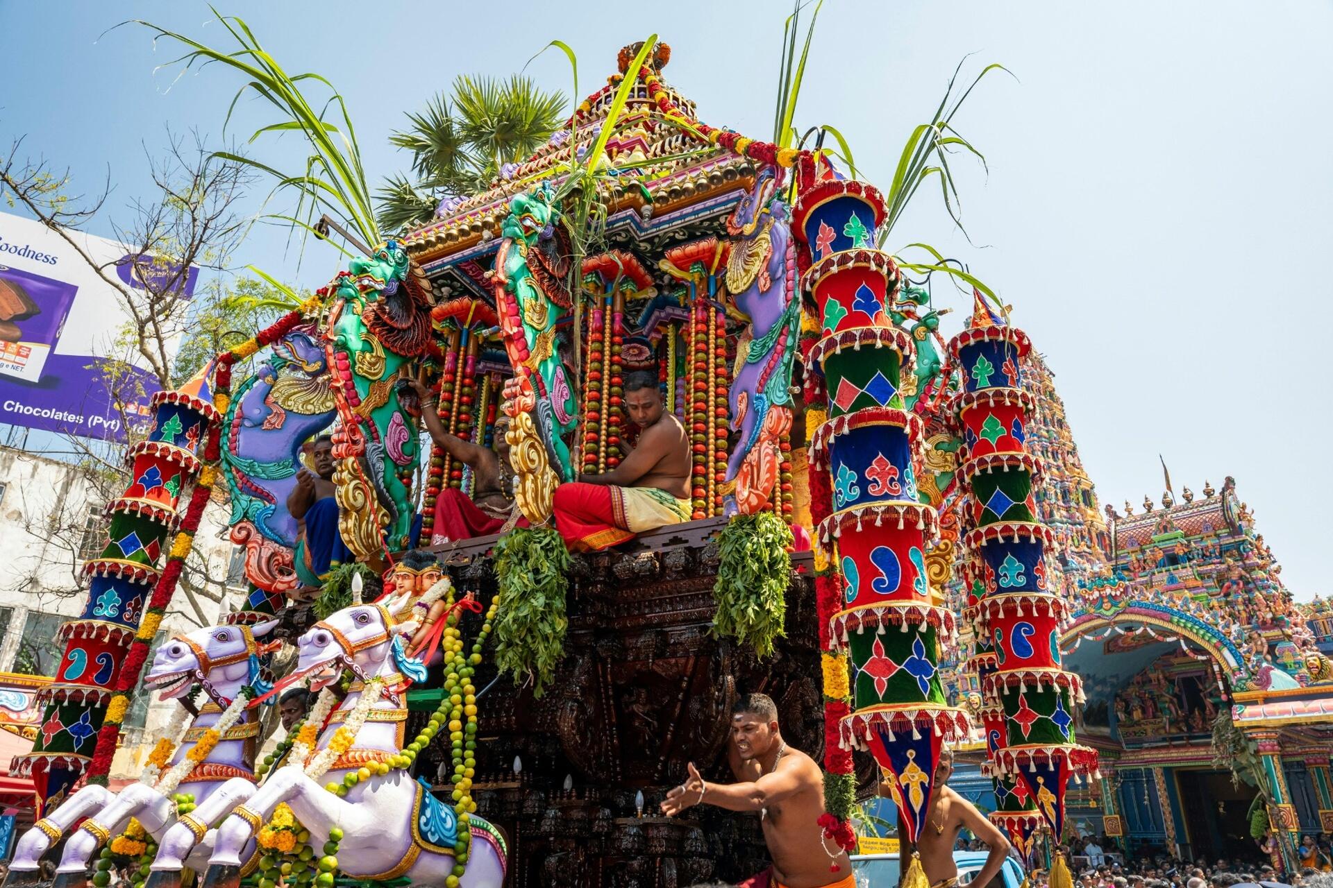 Colourful Tamil Hindu temple festival procession featuring an ornate ceremonial chariot, decorated figures, and devotees participating in a traditional religious celebration.
