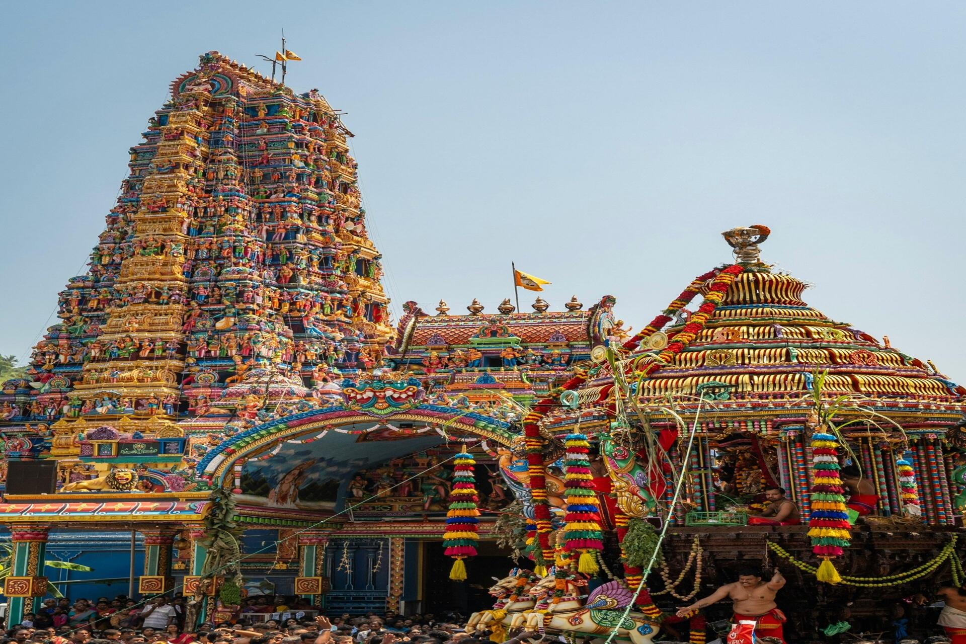 Colourful South Indian Hindu temple during a festival celebration, featuring ornate Dravidian architecture, decorated chariot structures, and gathered worshippers.