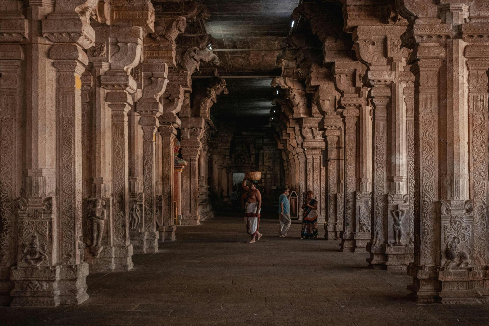 Interior corridor of a historic South Indian temple featuring intricately carved stone pillars, traditional Dravidian architectural detailing, and visitors walking through the sacred space.