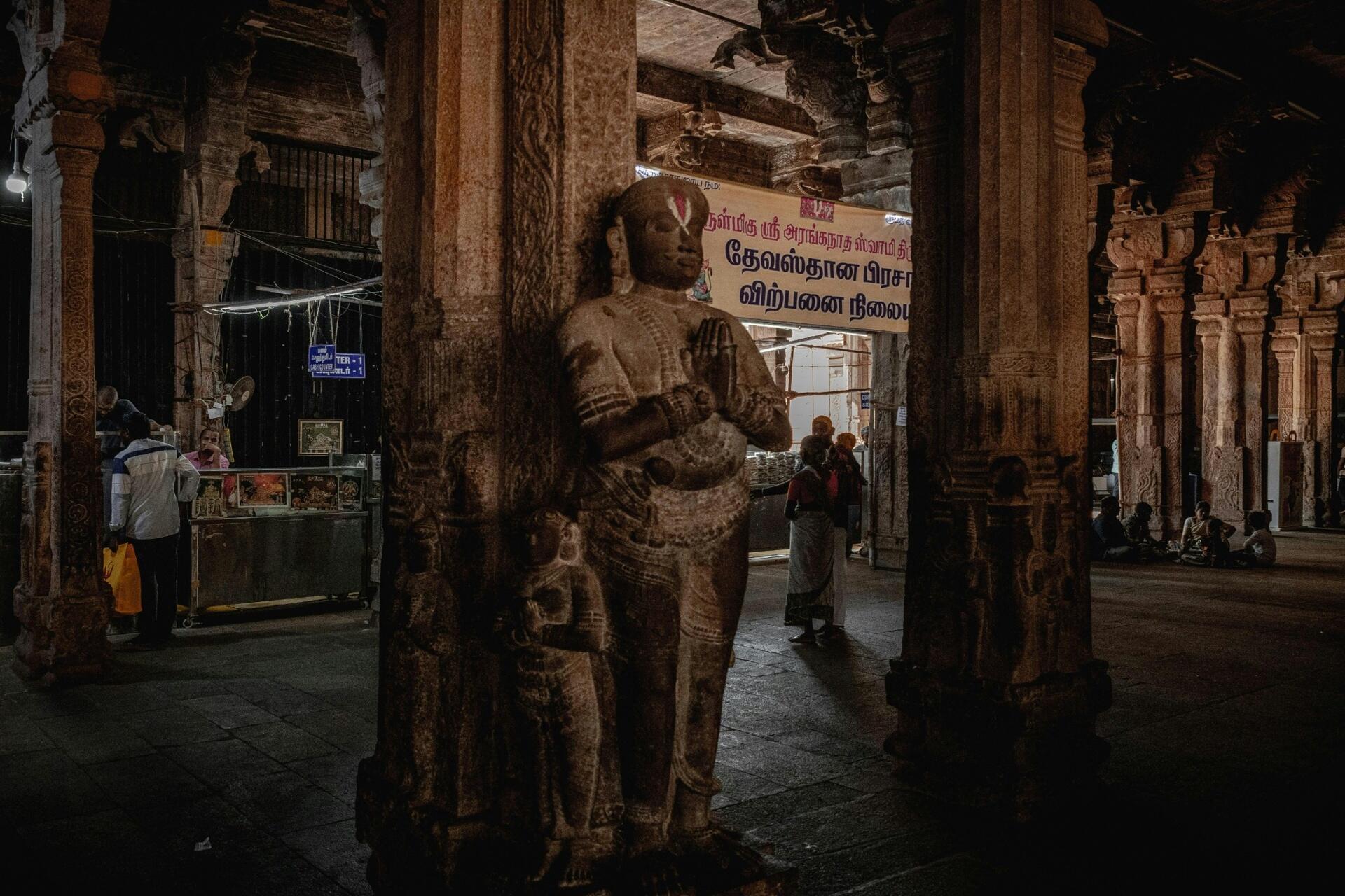 Interior of a historic South Indian temple featuring carved stone pillars, a sculpted temple figure, and traditional Tamil religious architectural details.