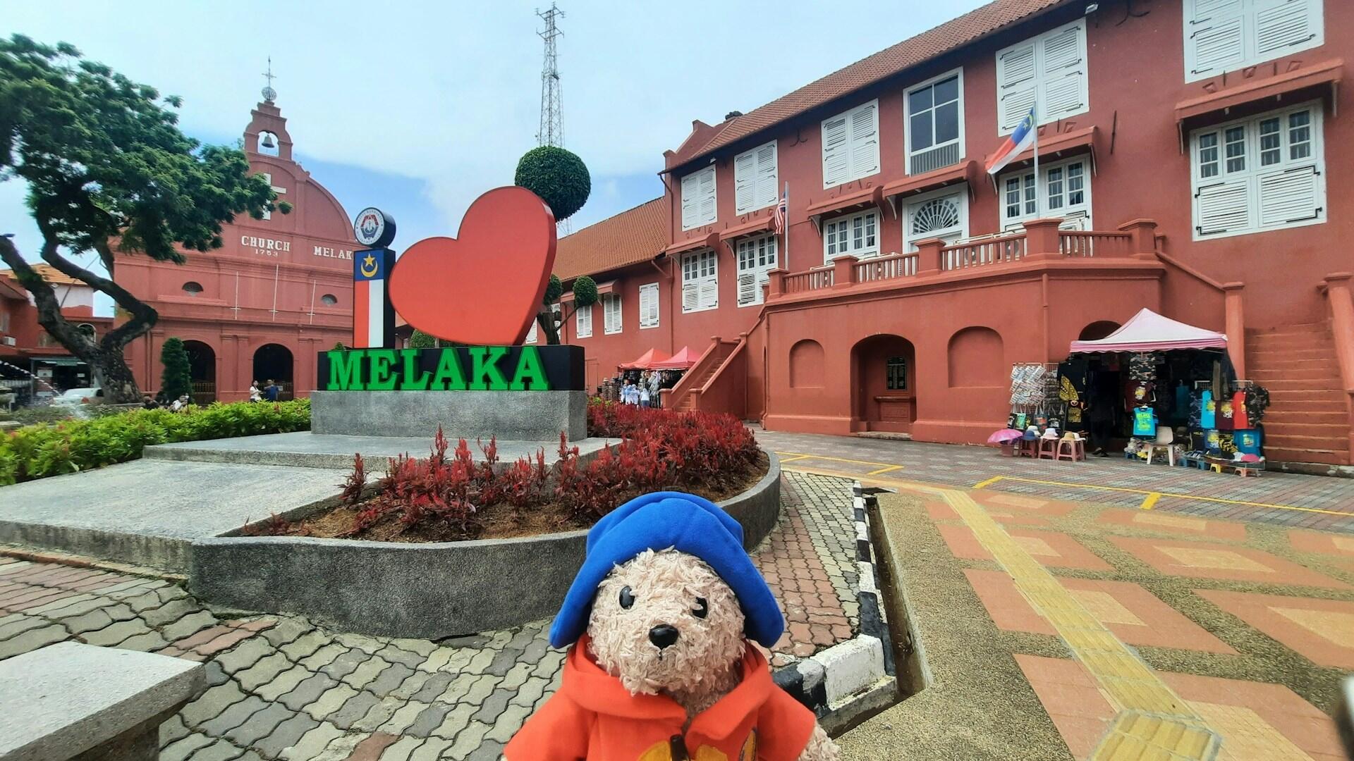 A teddy bear positioned in front of iconic Melaka landmarks.
