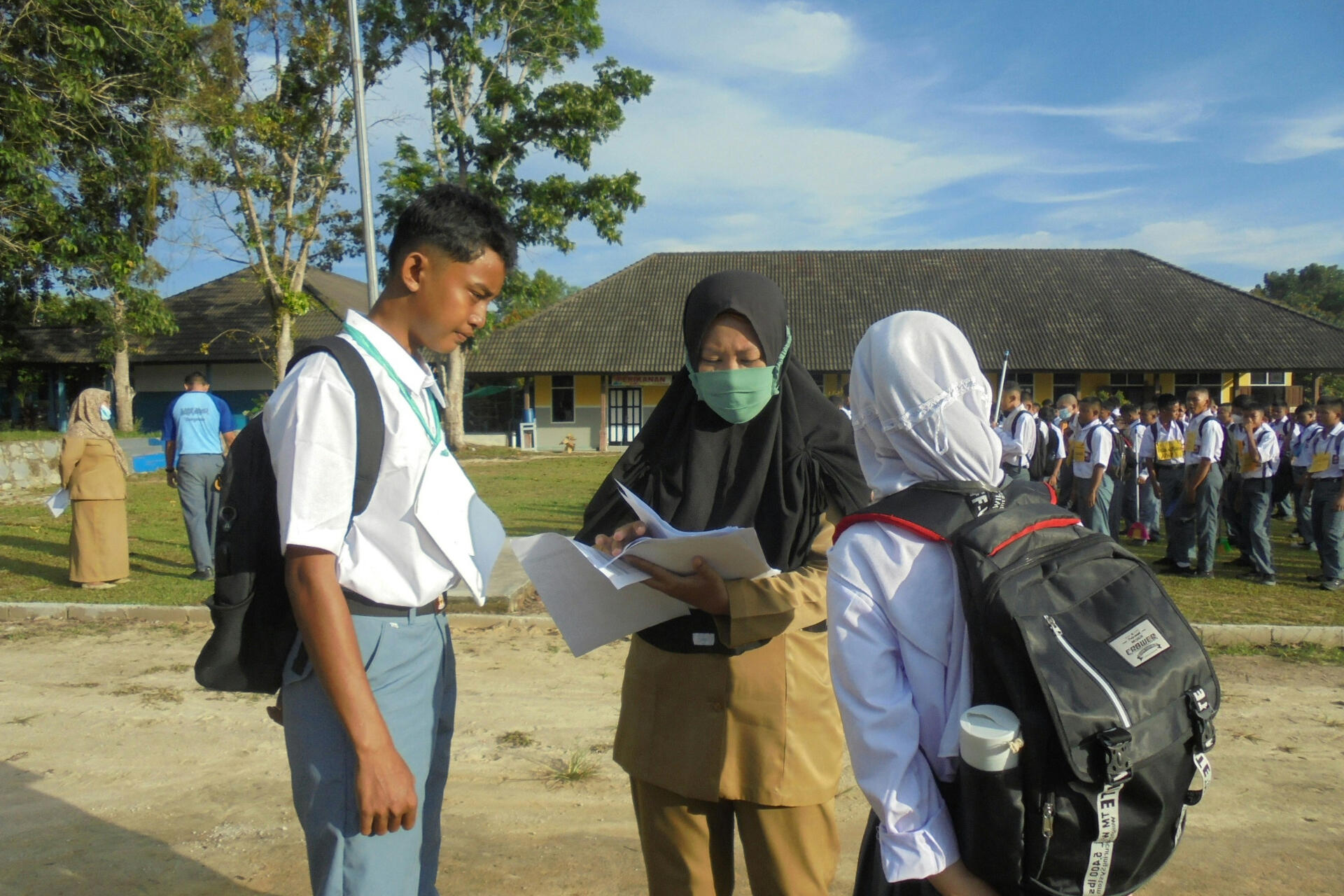 A teacher standing on the ground with a book and pen, tutoring two students who are standing side by side and attentively listening to the lesson.