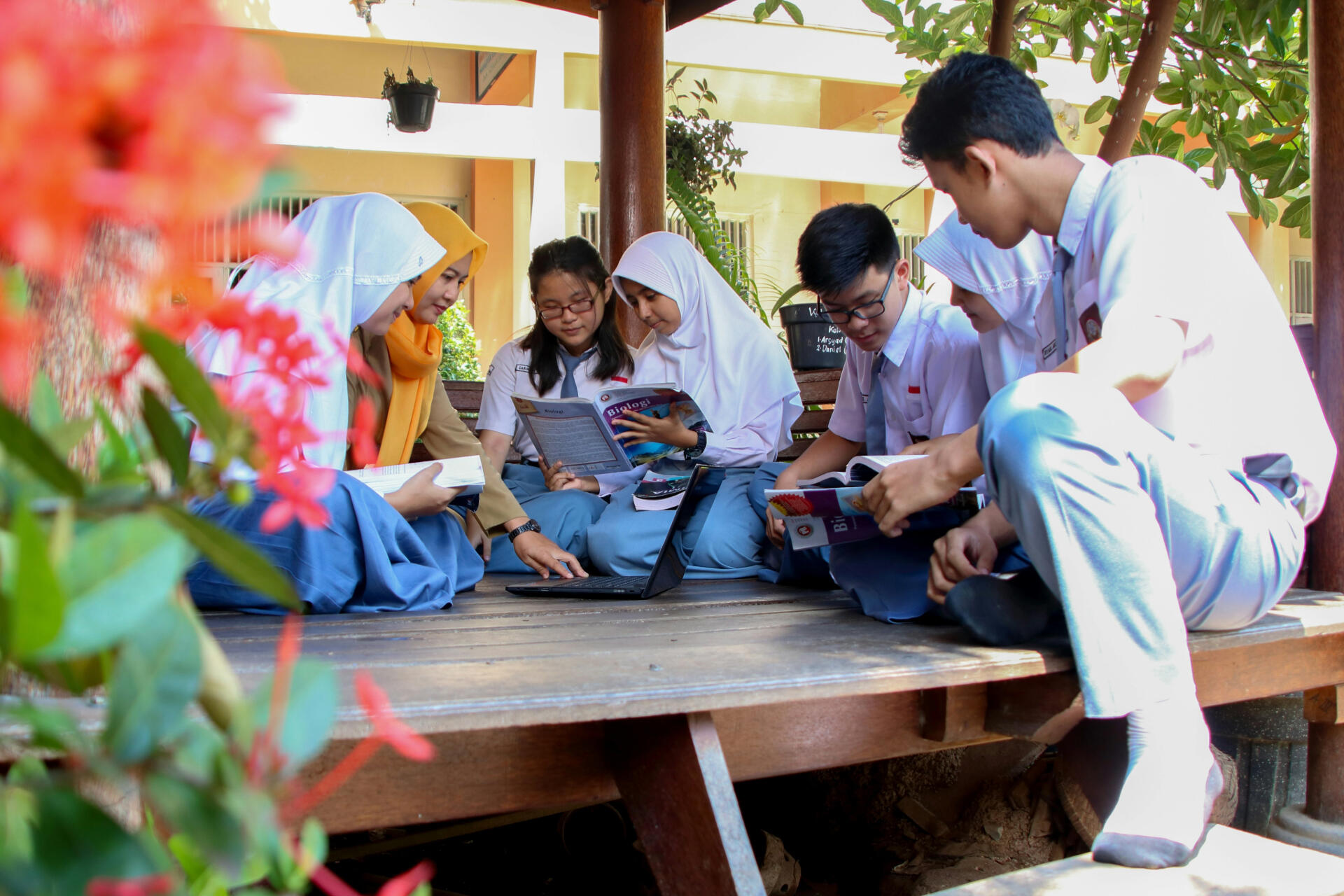 A group of high school students in uniforms sitting together outdoors, studying books and using a laptop.