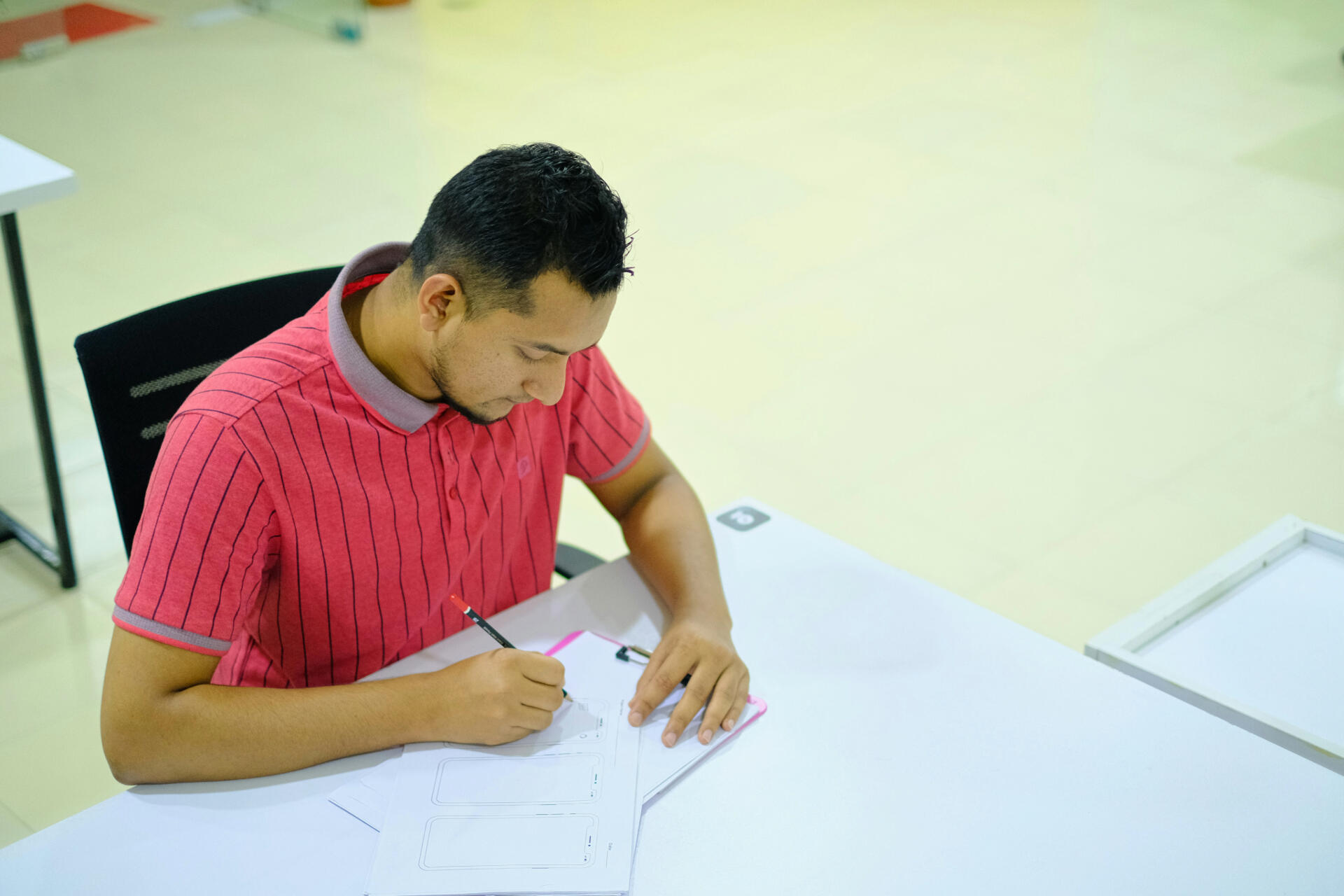 A young man in a red striped shirt sitting at a desk and writing notes on paper with a pencil.