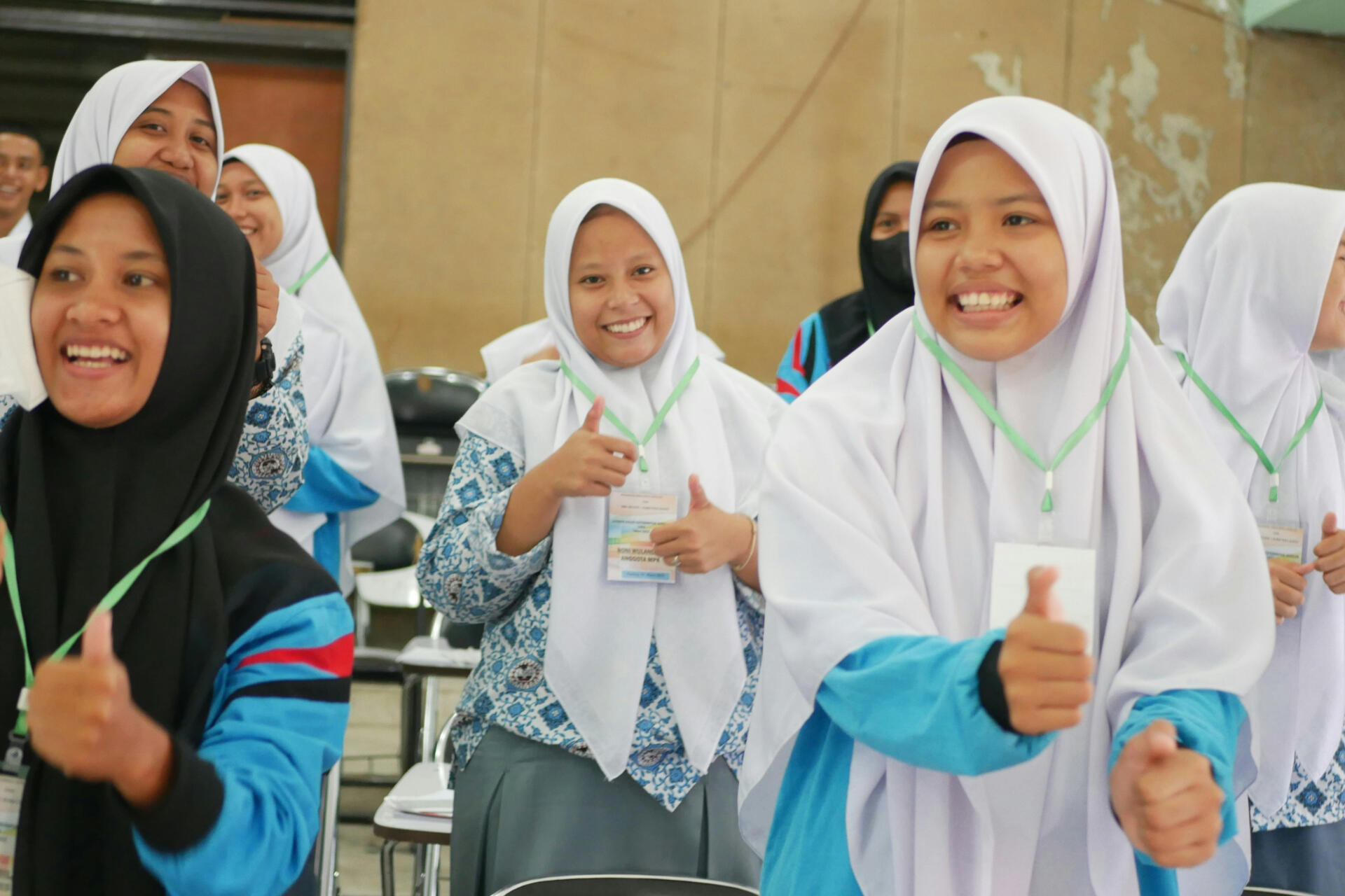 A group of smiling students wearing uniforms and hijabs, showing thumbs up in a classroom.