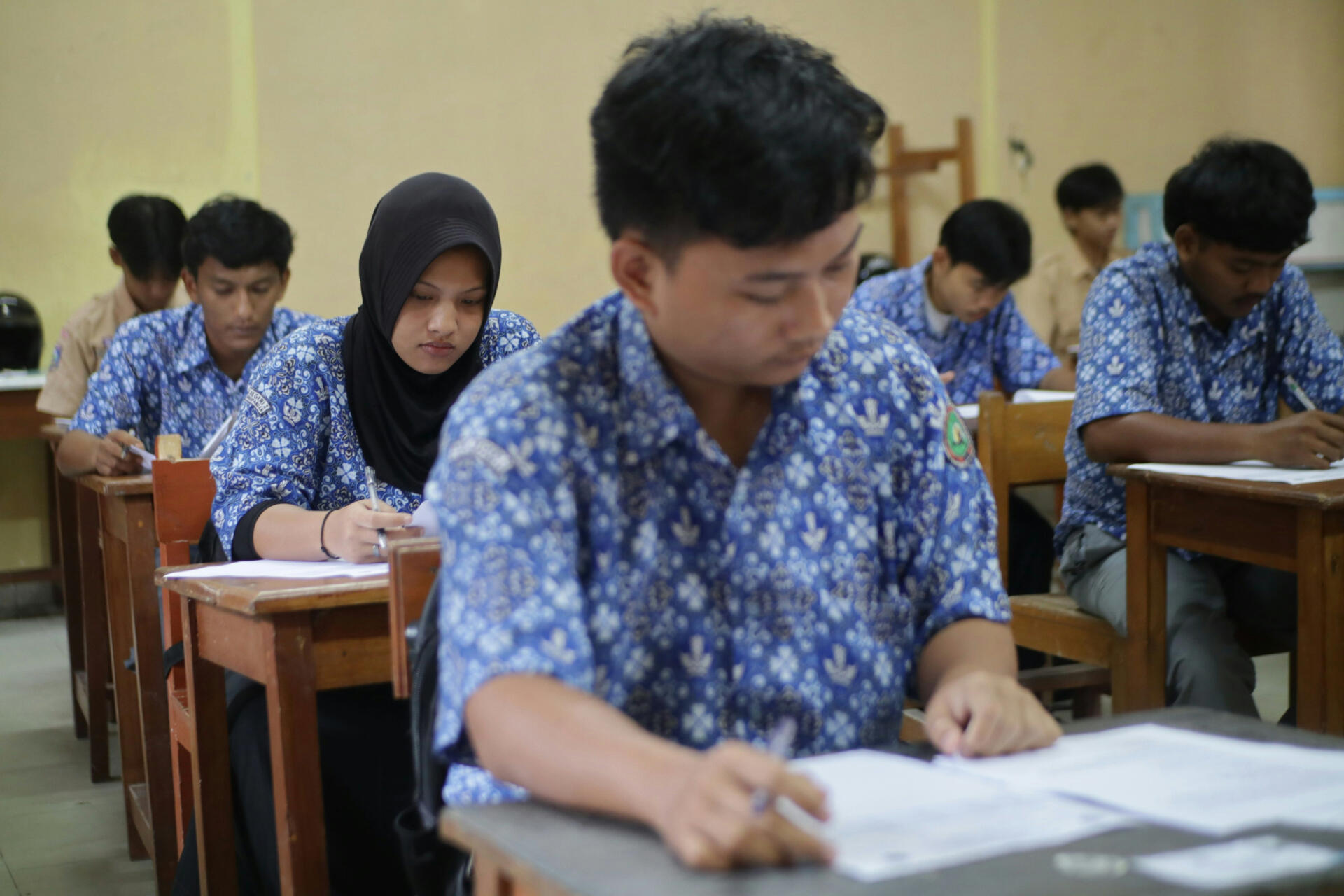 A group of students in uniform sitting in a classroom, writing answers during an examination.