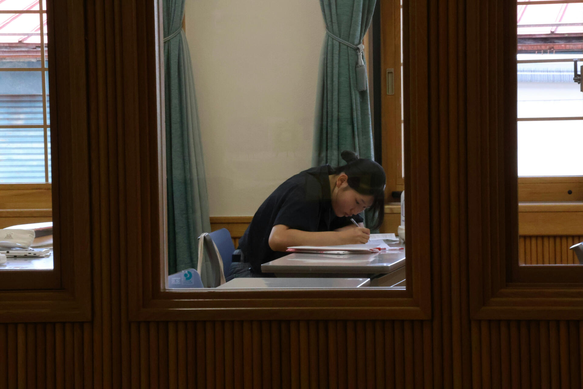 A student sitting at a desk in a classroom, focused on writing in a notebook.